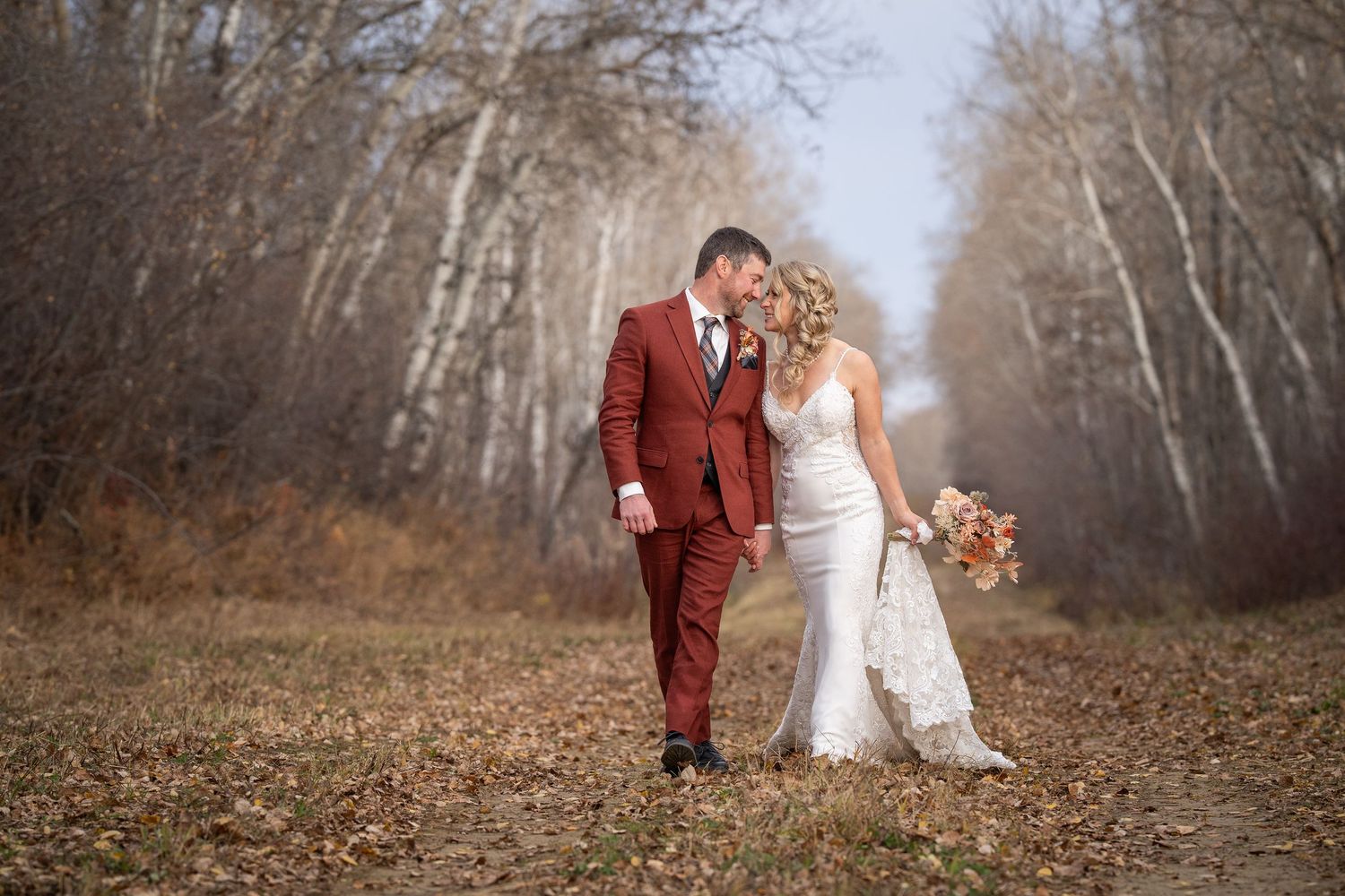 A couple in wedding attire walks along a misty autumn forest path lined with bare trees.