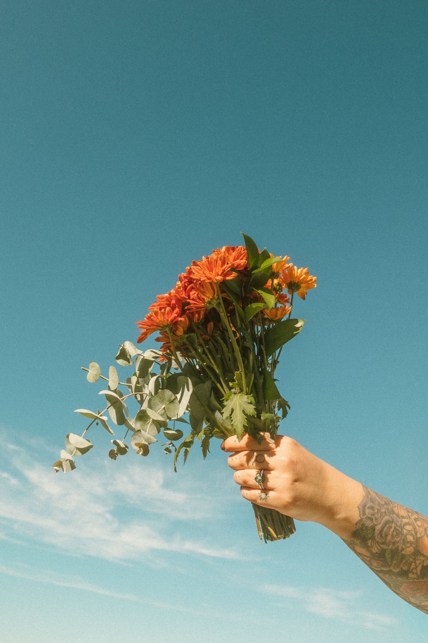 A tattooed arm extends upward holding a bouquet of orange and white flowers against a turquoise blue sky.