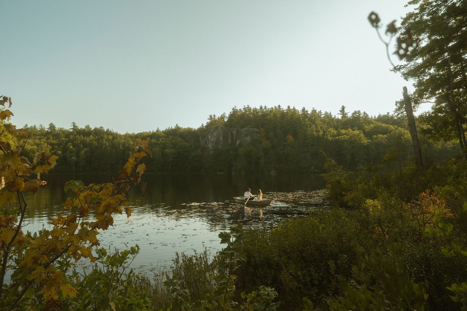 Scenic lake view surrounded by dense forest and rocky cliffs at sunset with golden light reflecting on calm waters.