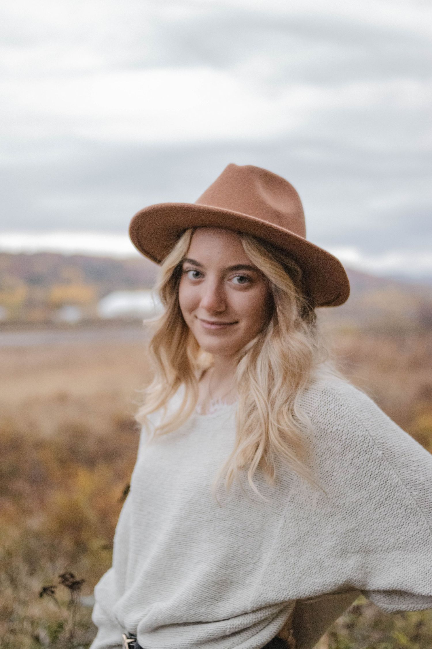 Person wearing a cozy white sweater and brown hat against an autumn landscape backdrop.