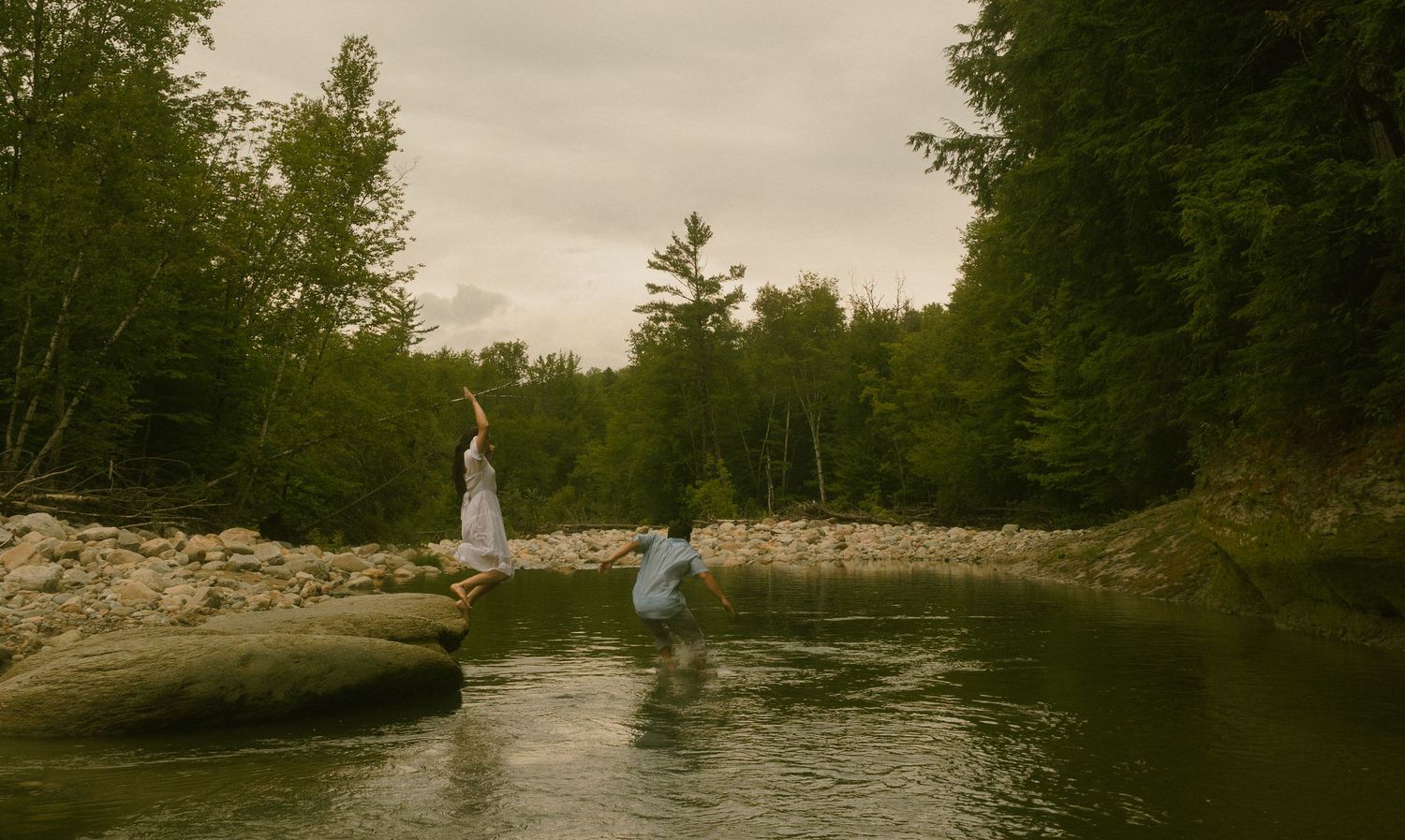 A serene river flows through a forested landscape at sunset with someone fishing from the rocky shoreline.