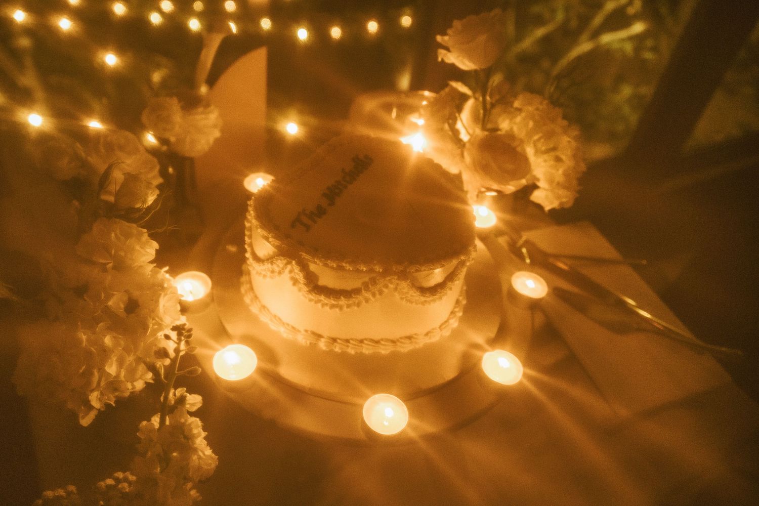 A romantic wedding cake illuminated by glowing tea lights and string lights creating a warm ambiance at night.