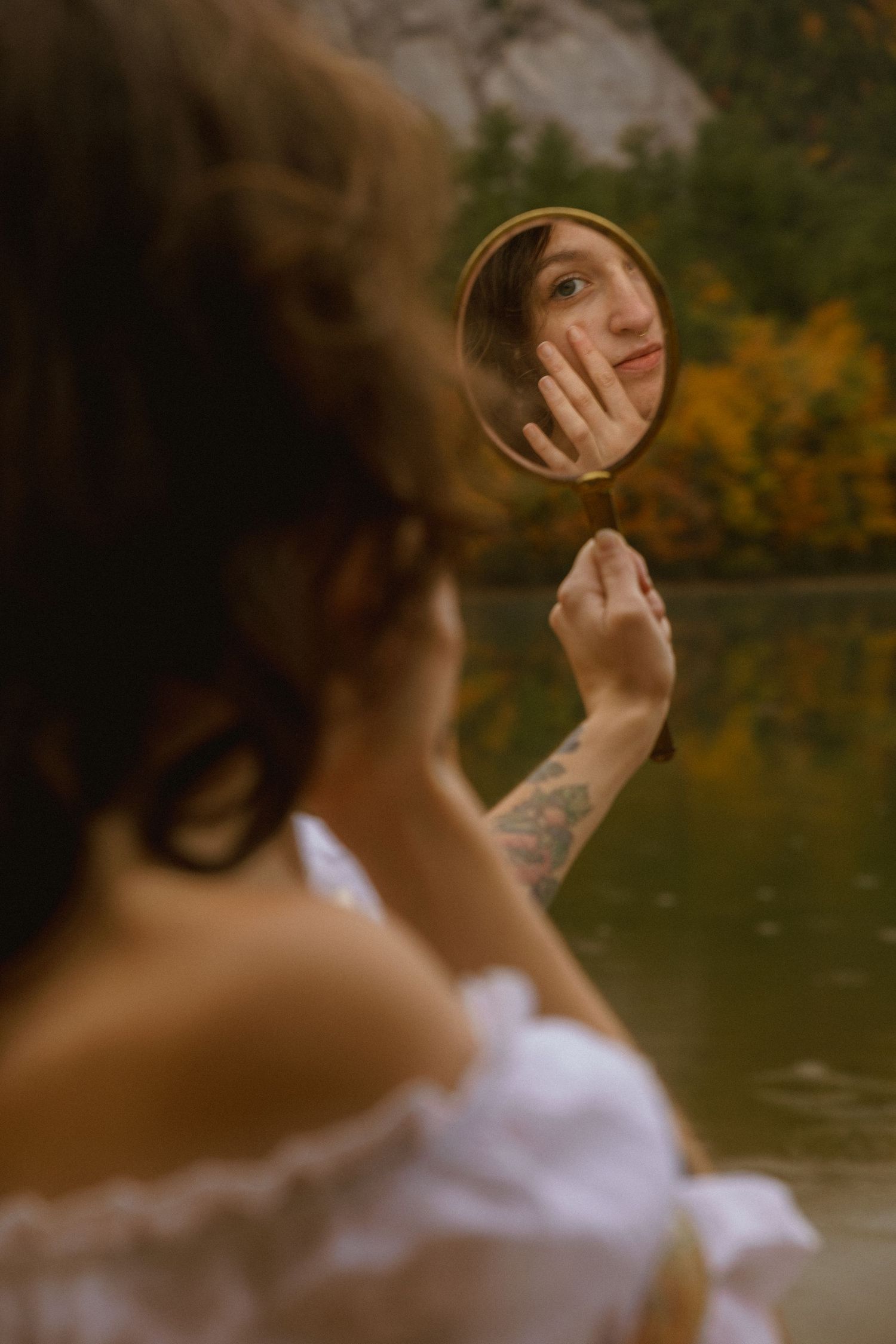 A person in a white top looks over their shoulder near a lake with autumn foliage in the background.