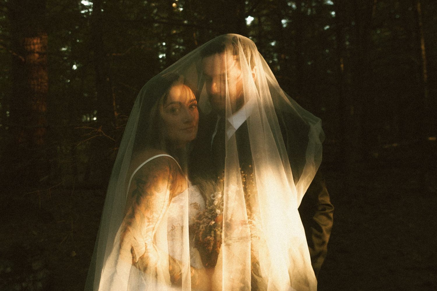 Ethereal bride in white dress and veil stands in dark forest creating a haunting, atmospheric wedding portrait.