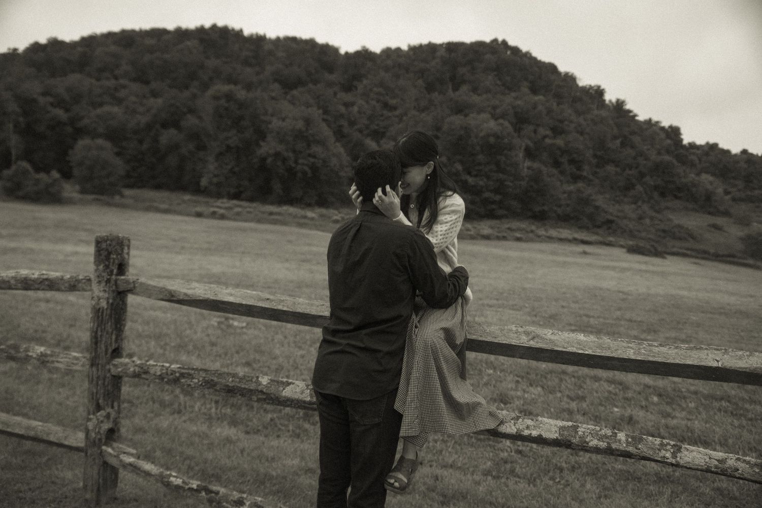 A vintage black and white photograph shows two figures standing by a wooden fence with rolling hills in the background.