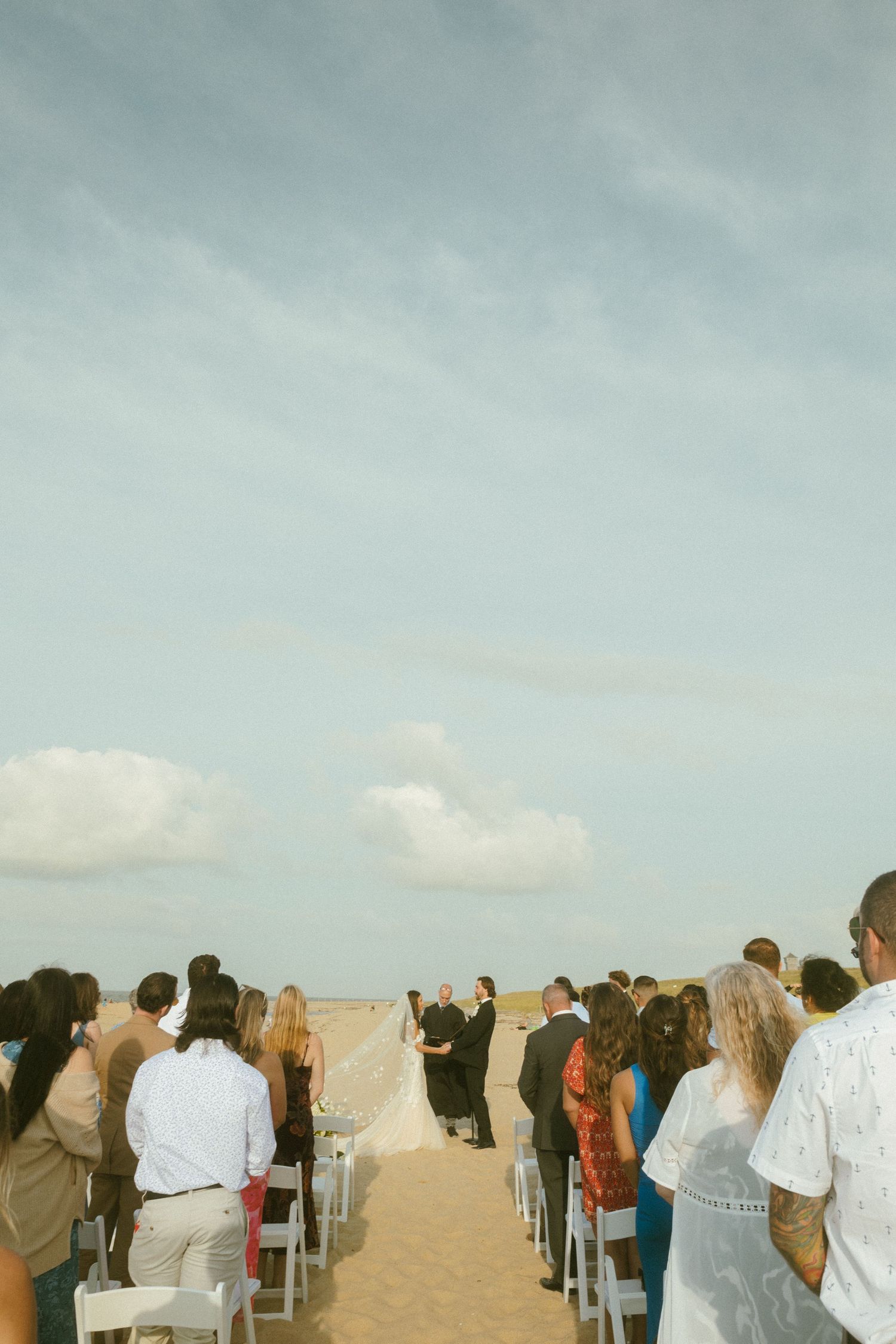 A crowd of wedding guests seated in white chairs on a sandy beach during an outdoor ceremony. Cape Cod wedding. beach Microwedding.