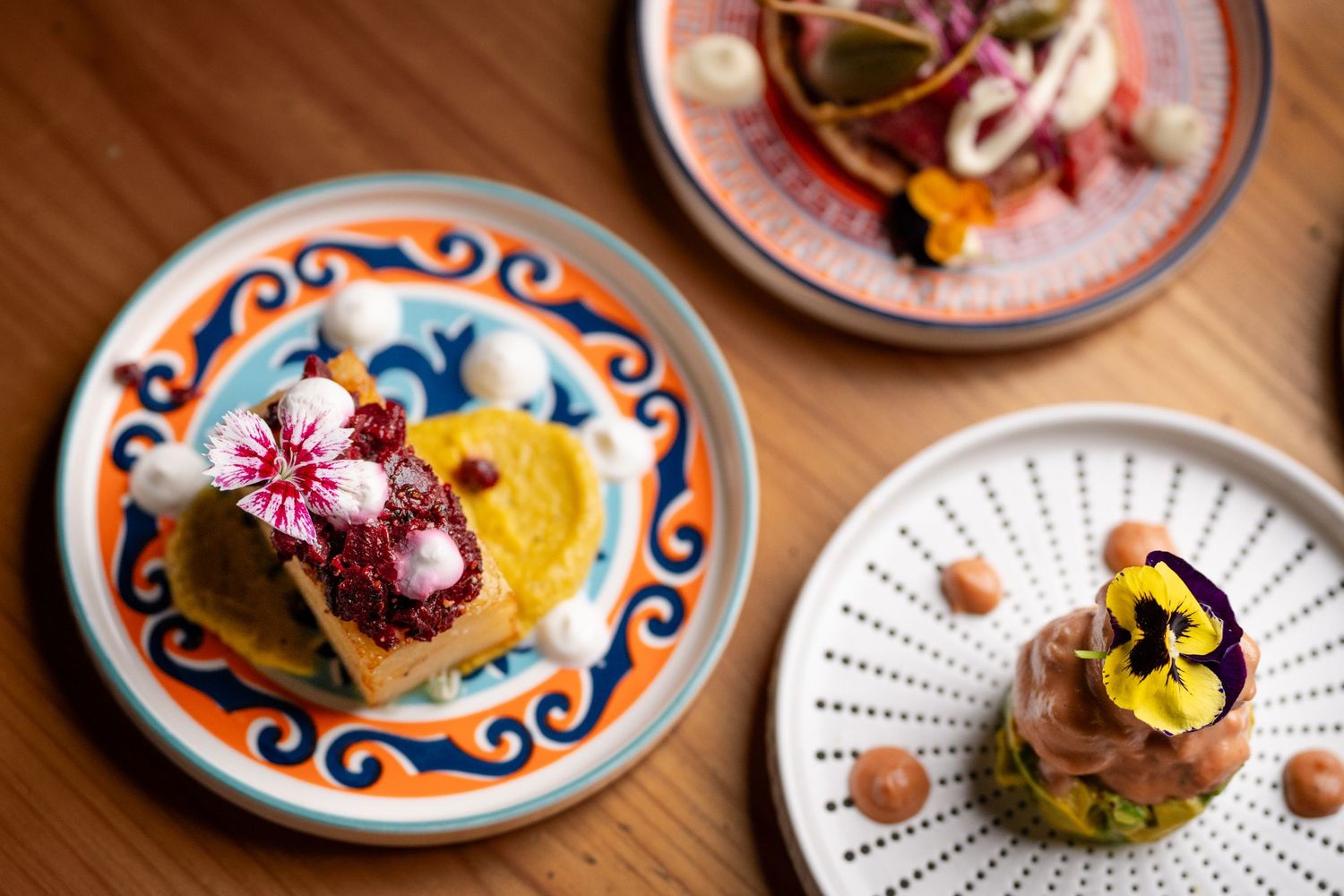 Three elegantly plated gourmet dishes on decorative plates, garnished with edible flowers on a wooden table.