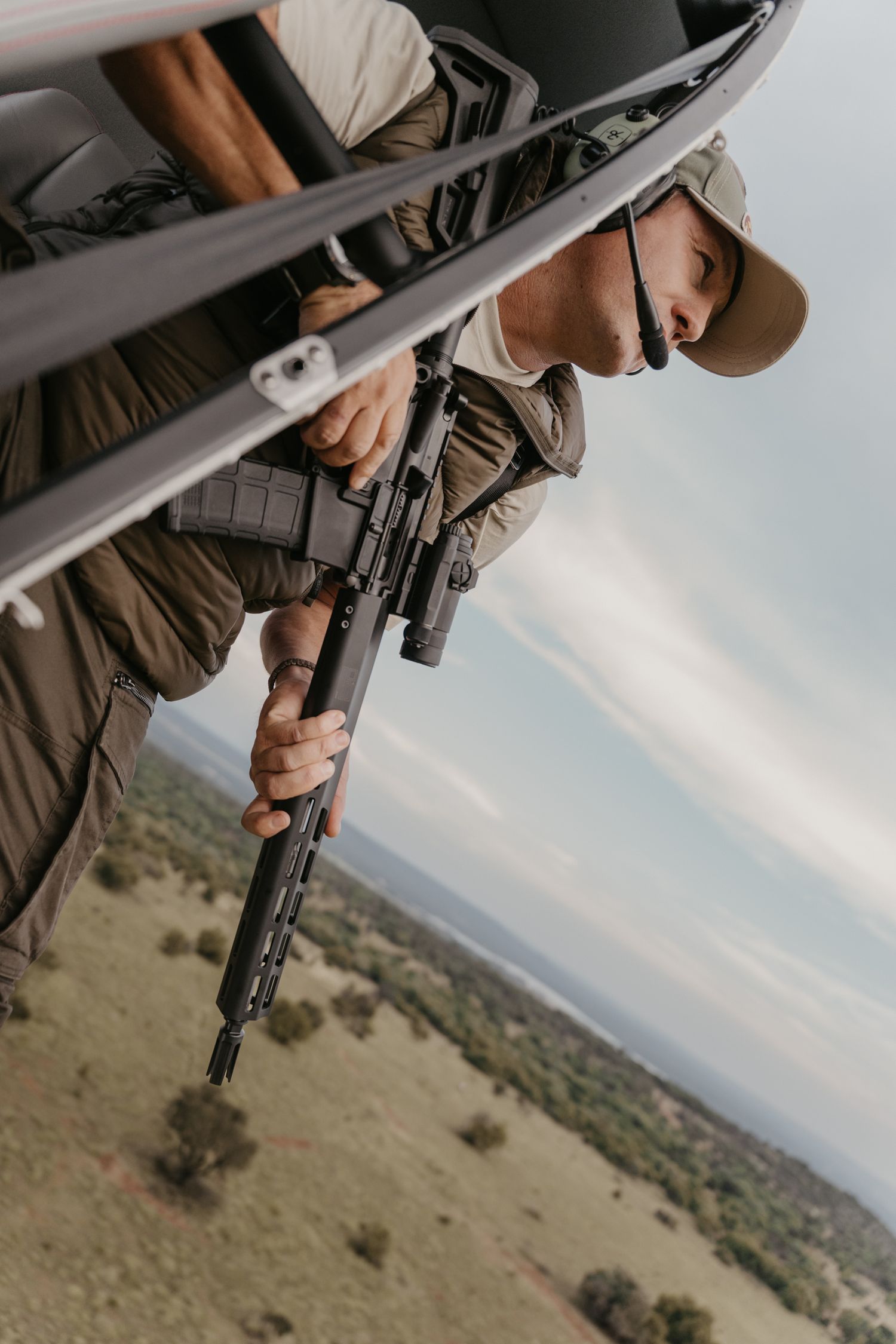 Armed ranger leaning out of a helicopter over African savanna landscape below.