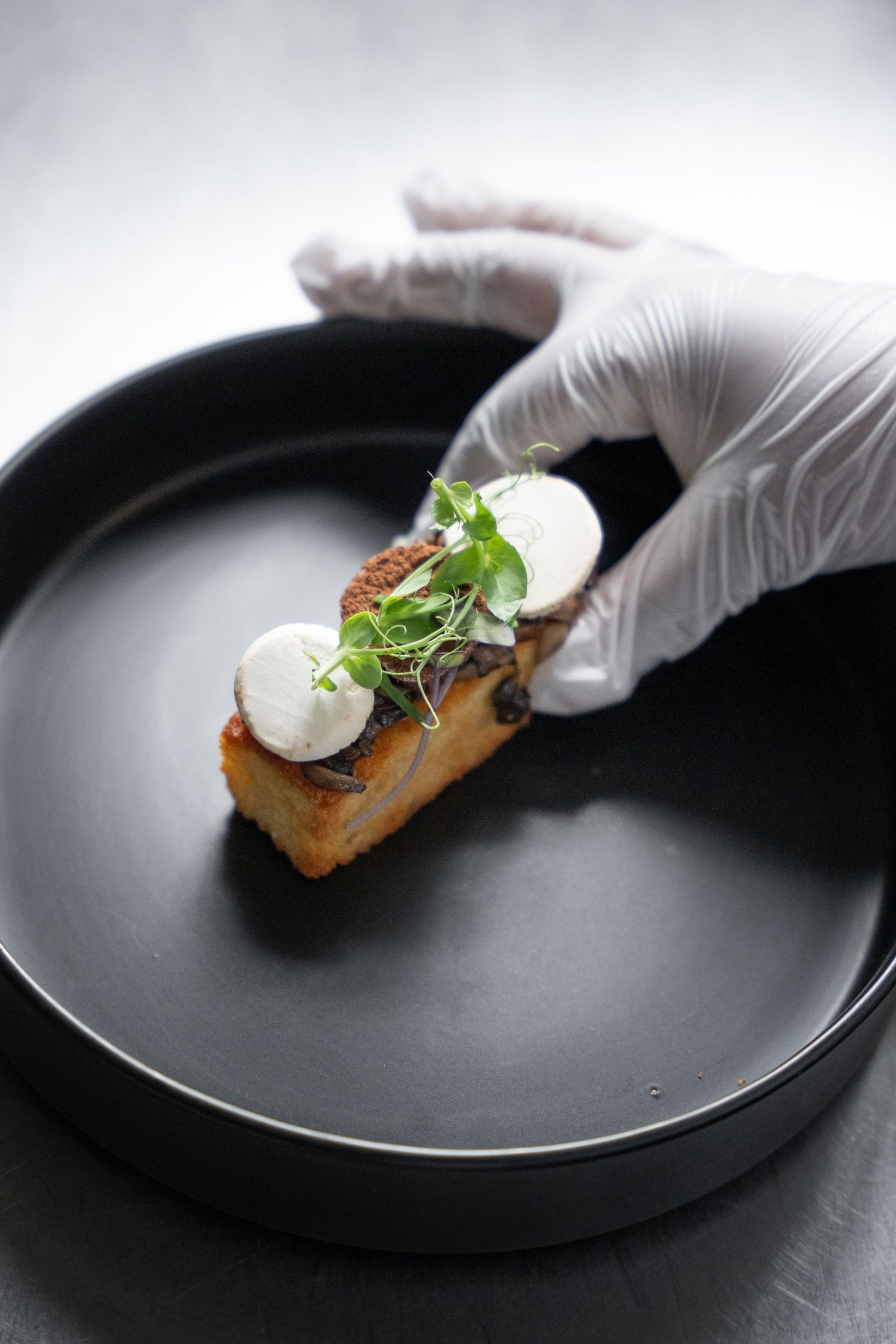 Chef in gloves placing artisan mushroom toast with goat cheese rounds and microgreens on black plate.