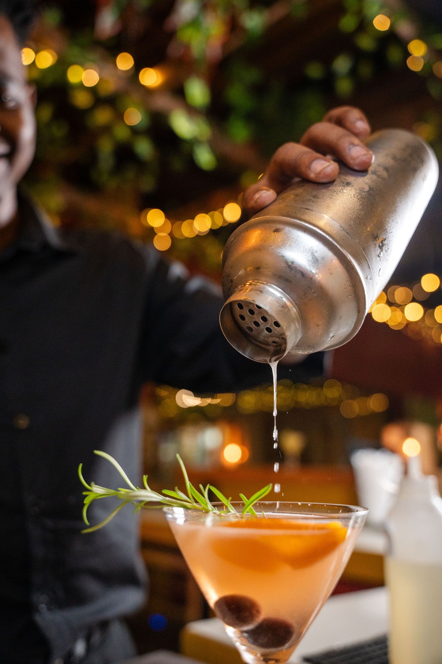 Bartender pouring a cocktail from a shaker into a martini glass garnished with rosemary and olives.