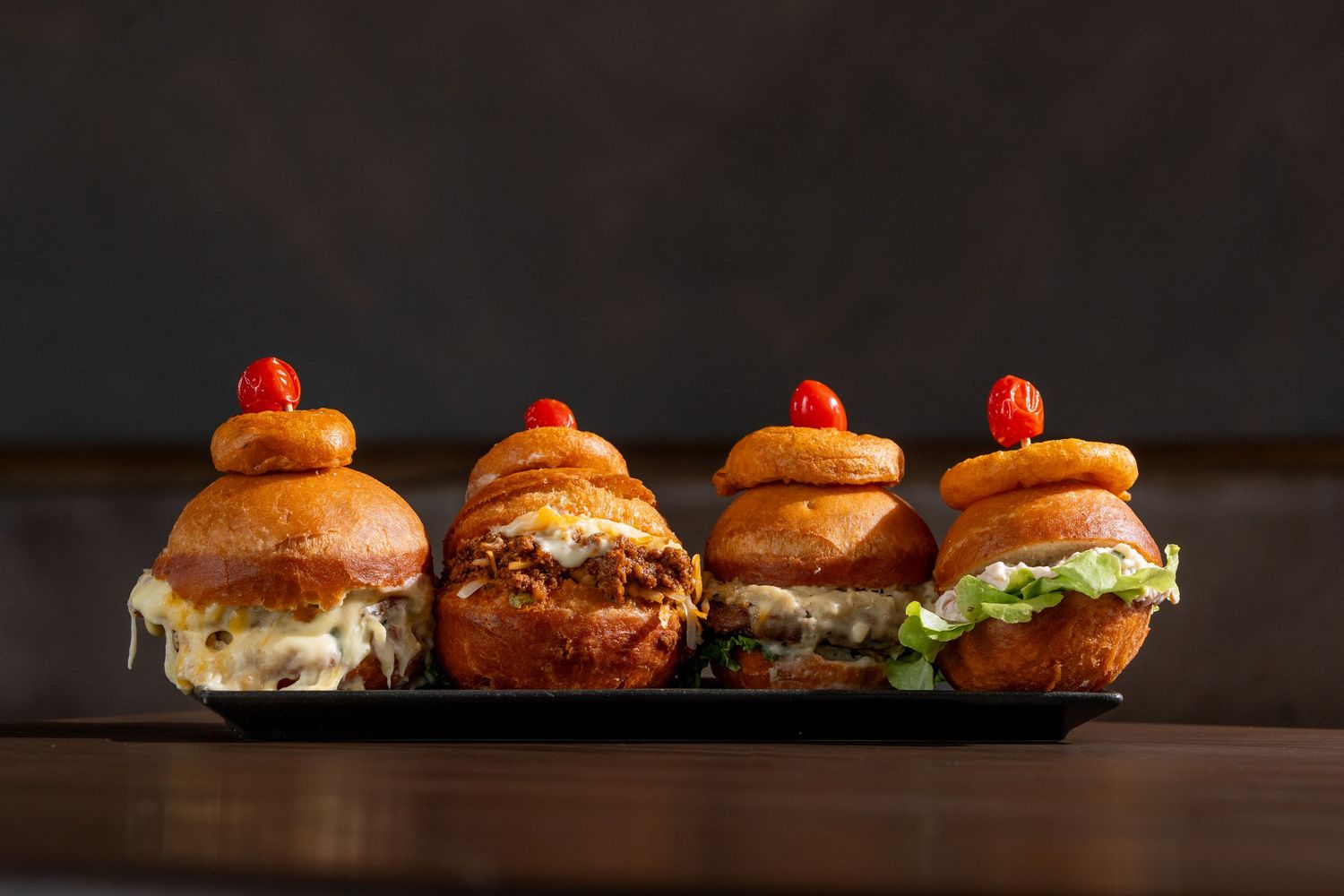 Four gourmet mini sliders topped with onion rings and cherry tomatoes served on a dark rectangular platter.