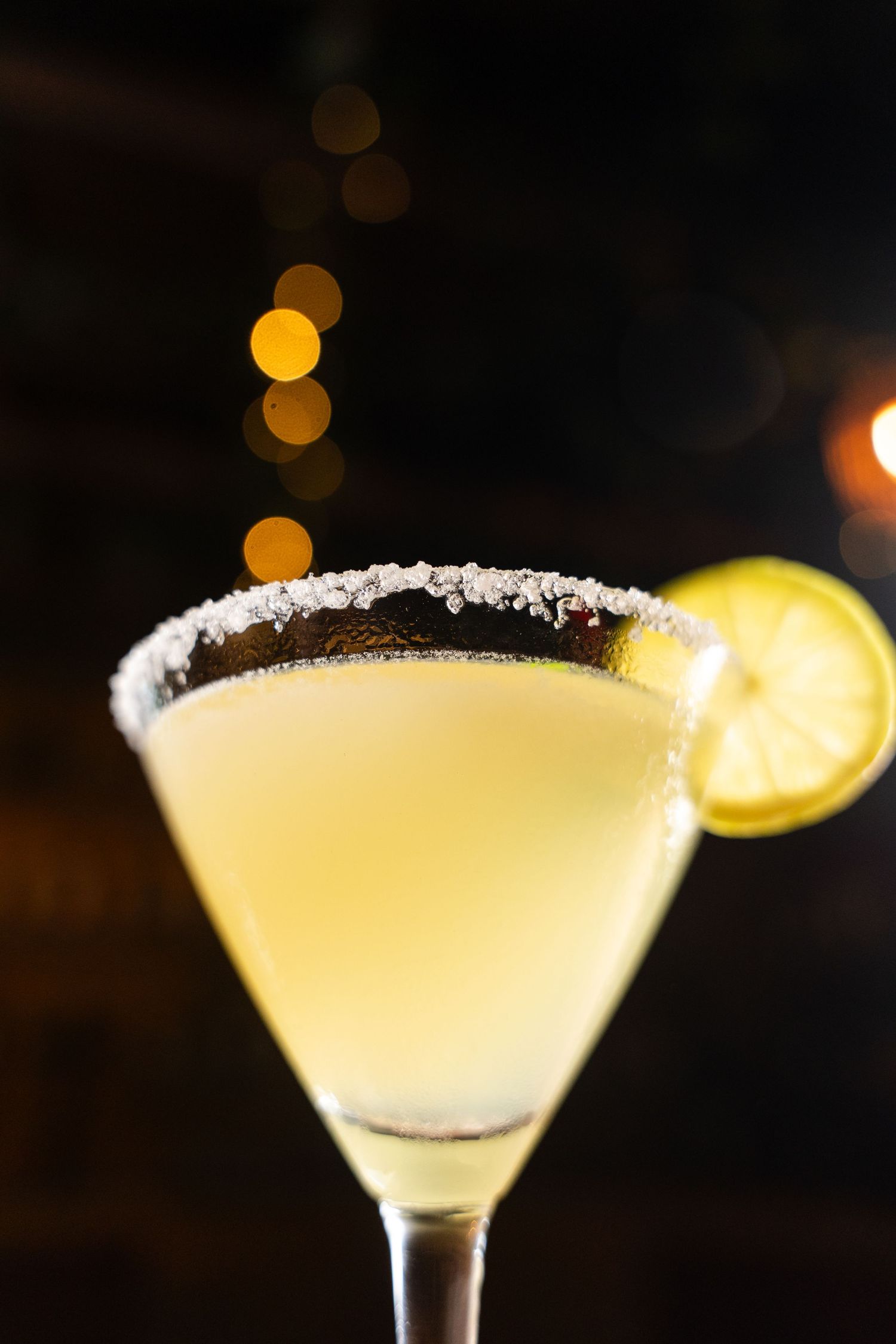 A classic margarita cocktail in a salted rim martini glass garnished with a lime slice against a dark bokeh background.