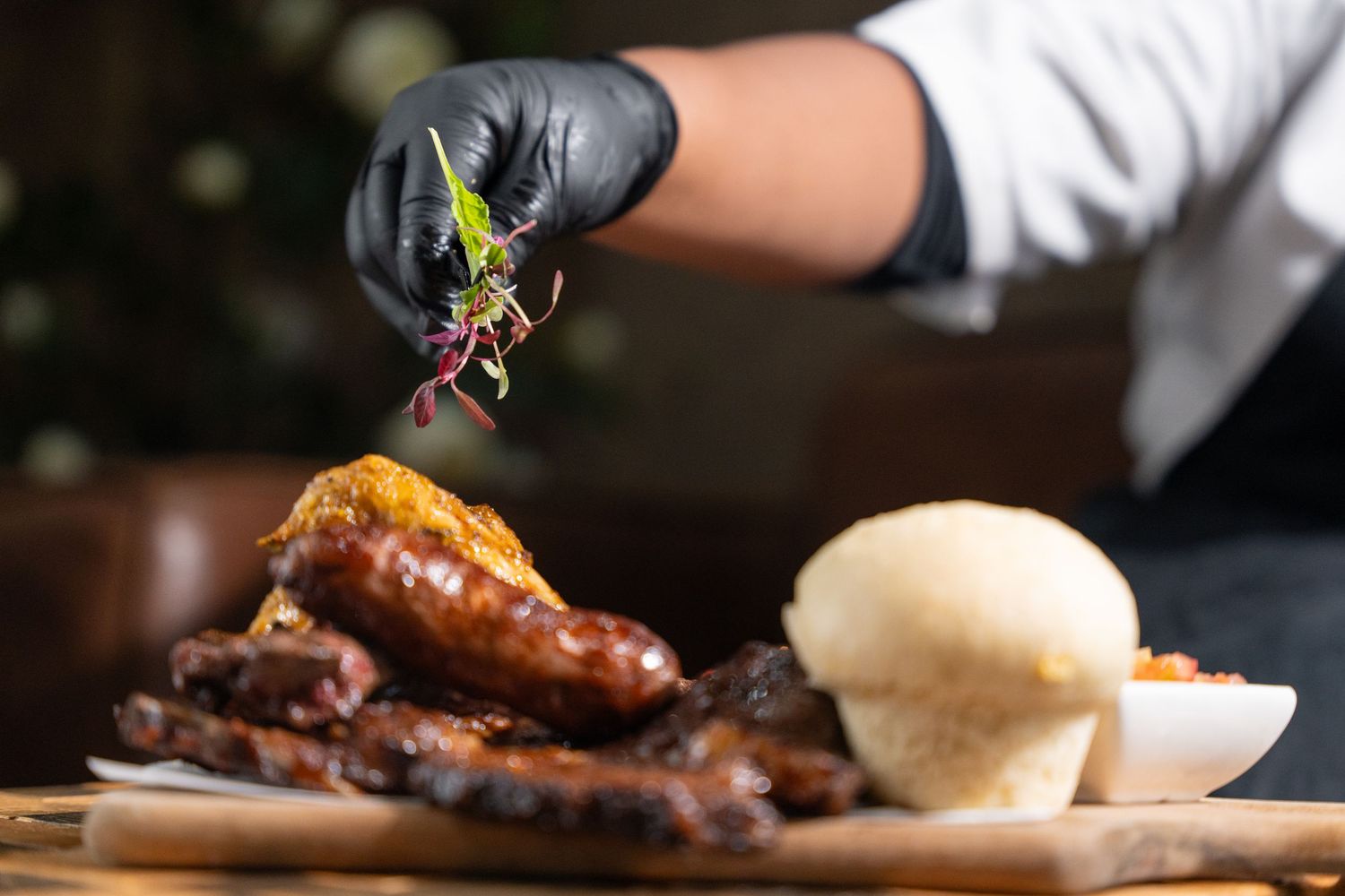 Chef garnishing a platter of grilled sausages, ribs, and a bread roll with fresh herbs using black gloves.