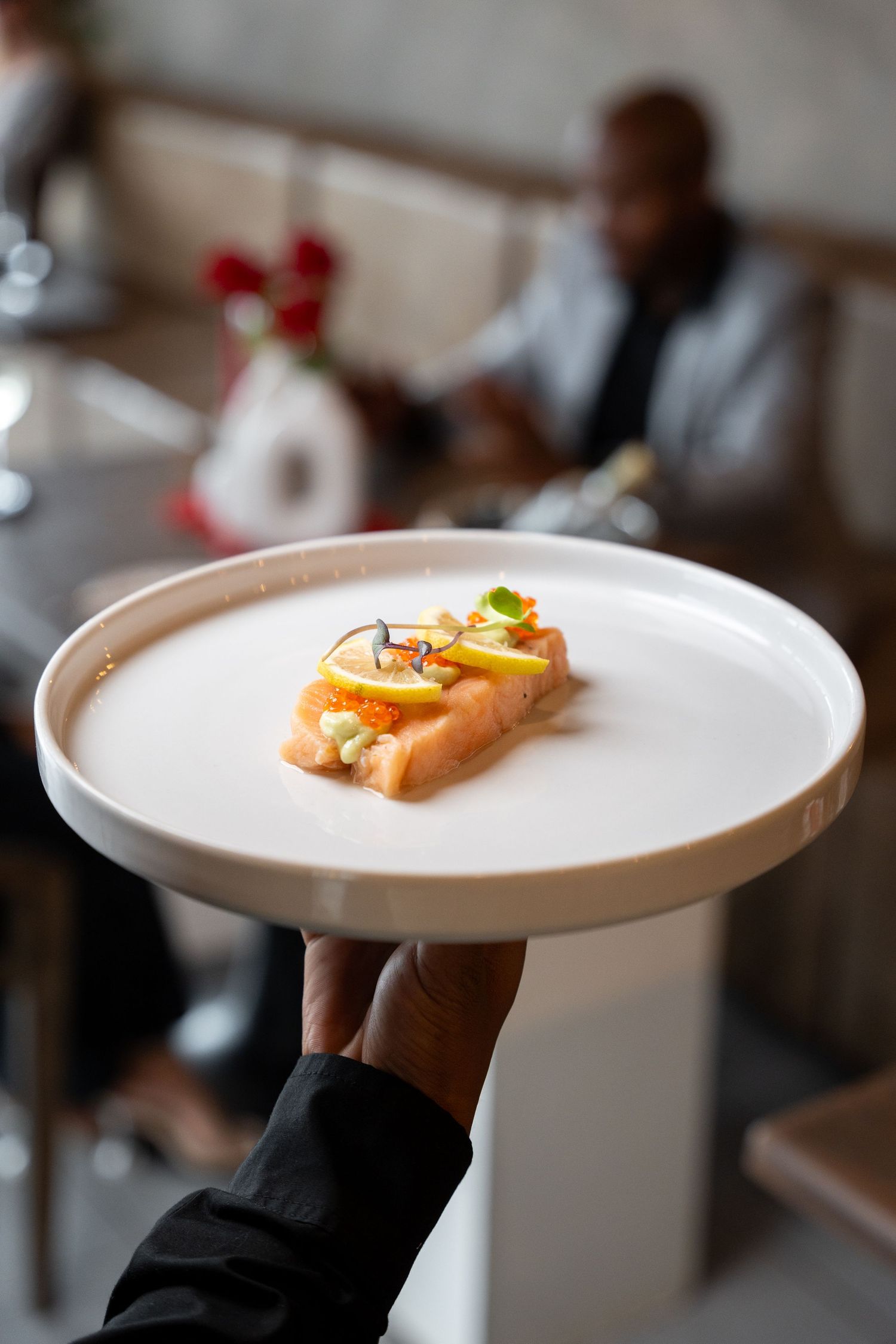 Waiter holding a white plate with an elegantly plated salmon dish garnished with lemon slices in a upscale restaurant.