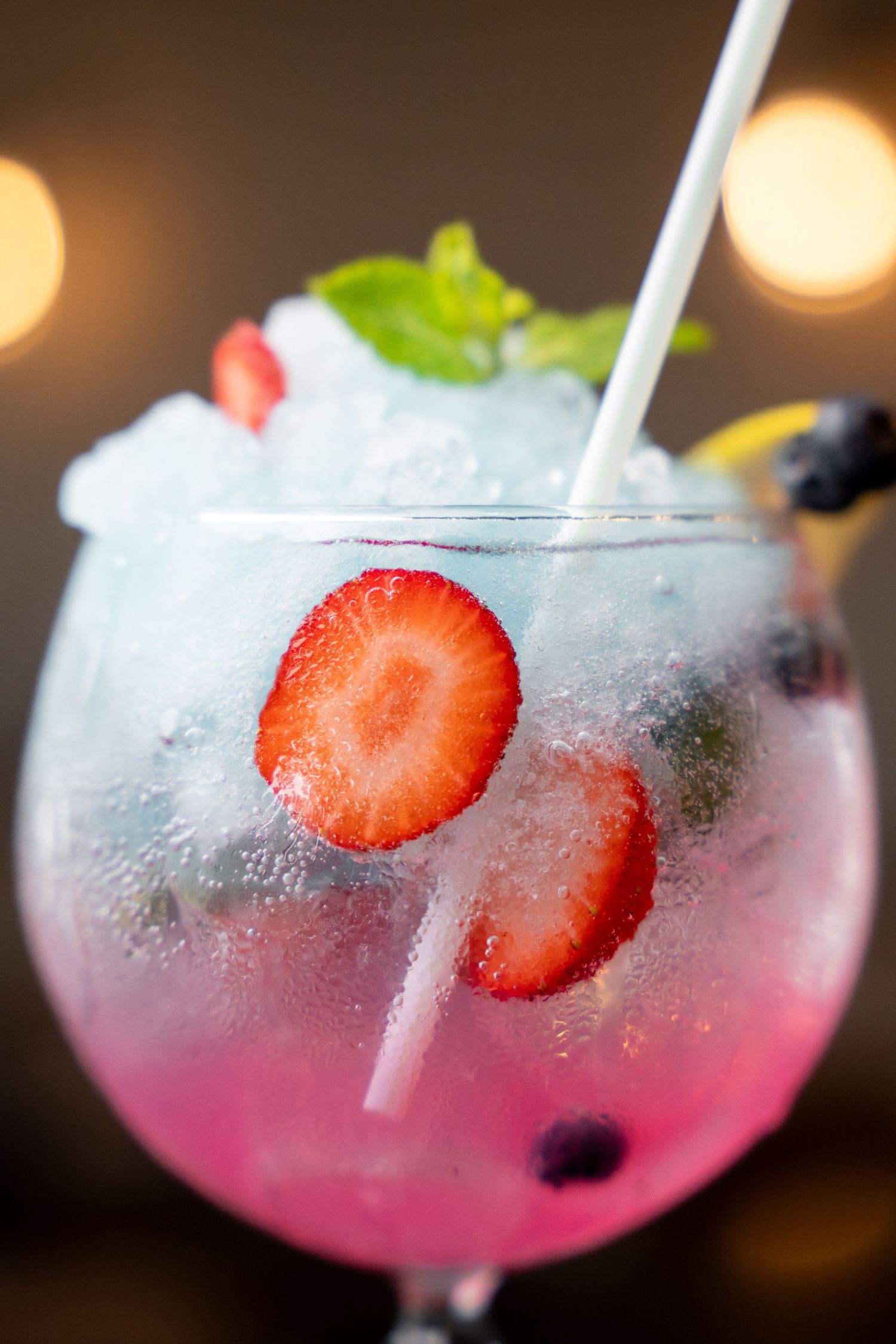A vibrant pink and blue cocktail with crushed ice, fresh strawberries, blueberries, mint, and a straw in a balloon glass.