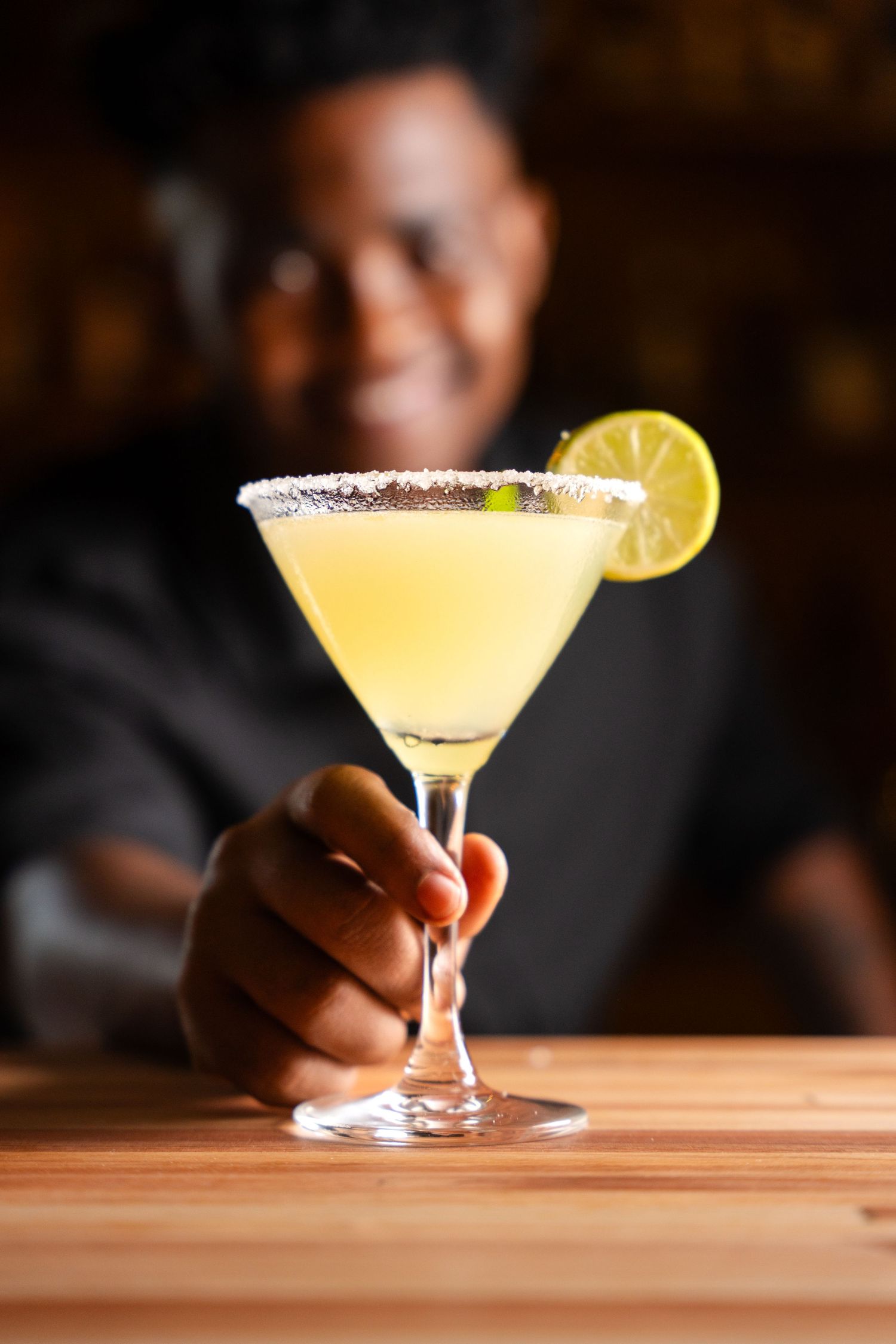 Bartender serving a yellow margarita cocktail in a salted rim martini glass garnished with a lime slice.