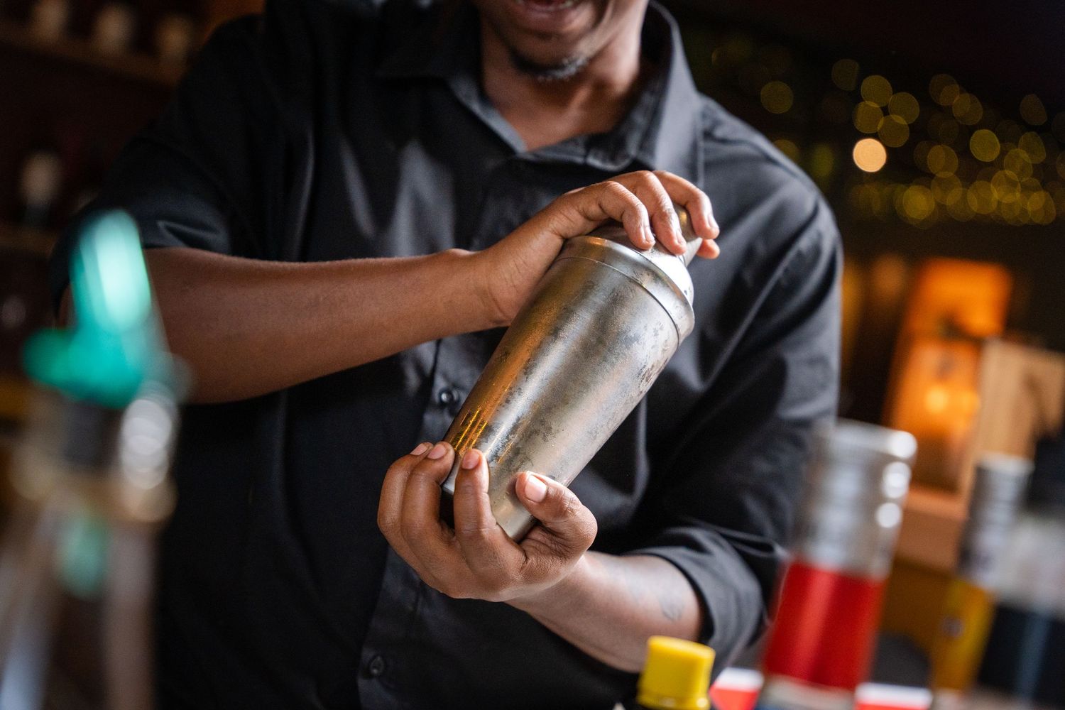 Bartender shaking a cocktail in a stainless steel shaker at a bar with bokeh lights in background.