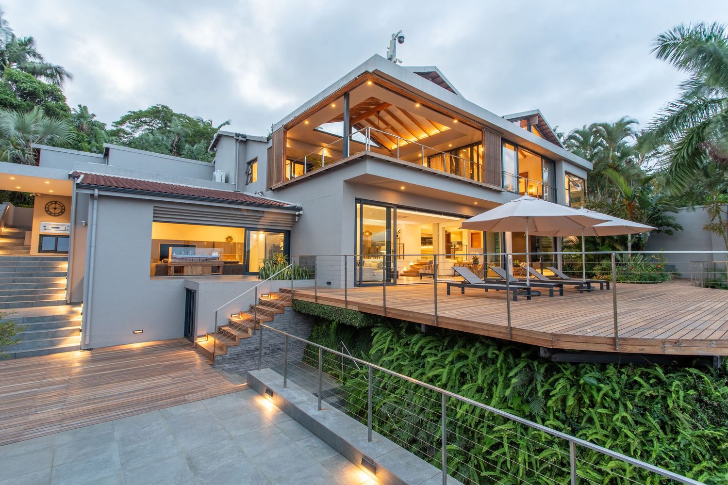 Luxury modern home with wooden deck, glass railings, lush tropical landscaping, and warm interior lighting at dusk.
