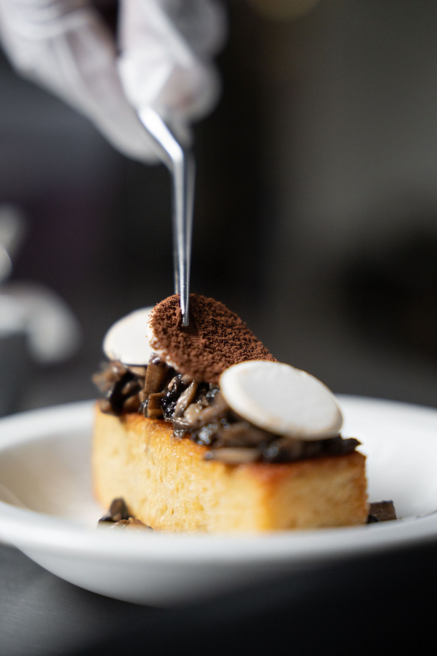 Chef plating gourmet mushroom bruschetta on toasted bread with white chocolate discs and cocoa crumble on white plate.