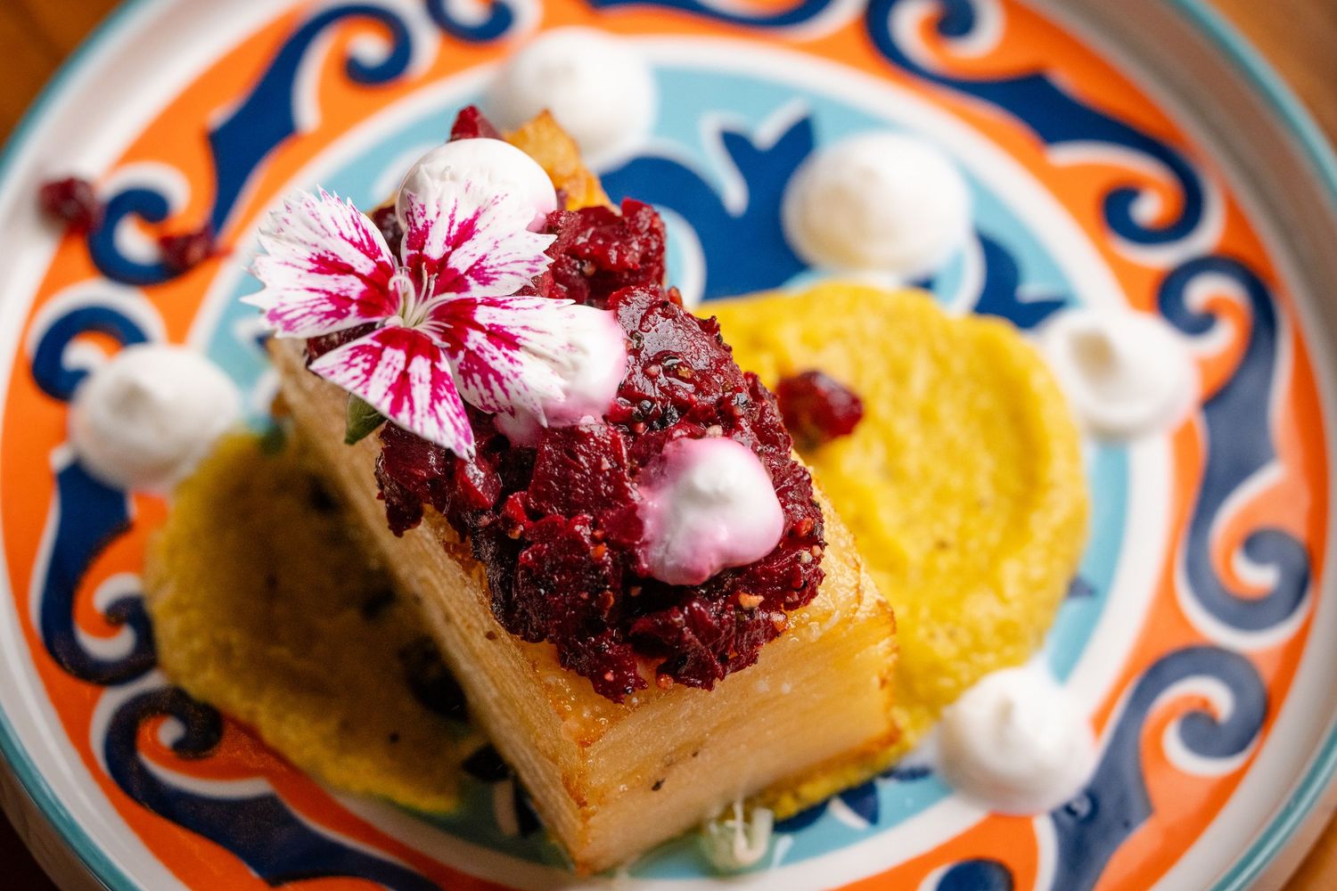 Elegant dessert with raspberry compote, edible flower, and cream on golden cake, served on colorful ceramic plate.