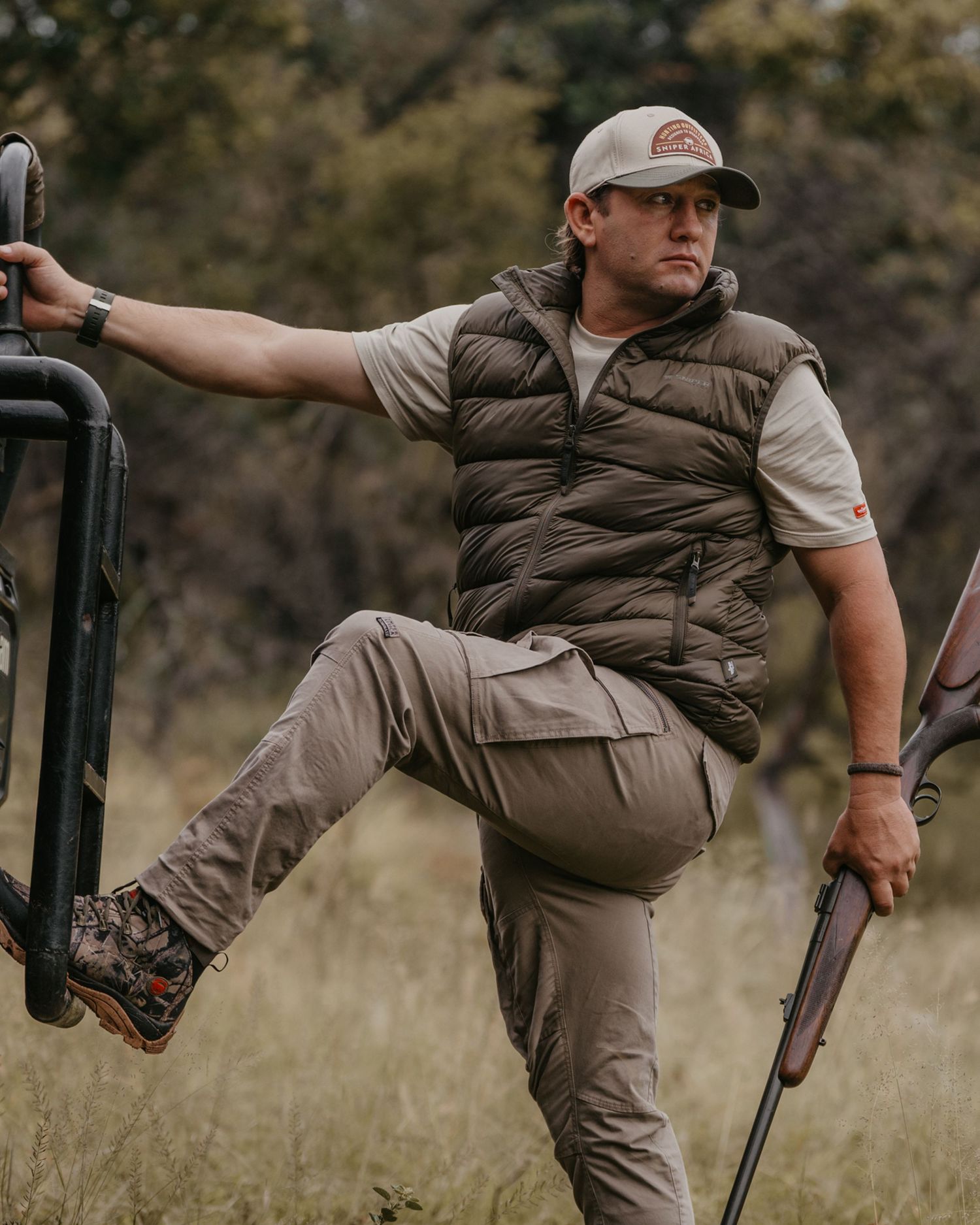 Man in olive puffer vest holding rifle while stepping onto safari vehicle in bush.