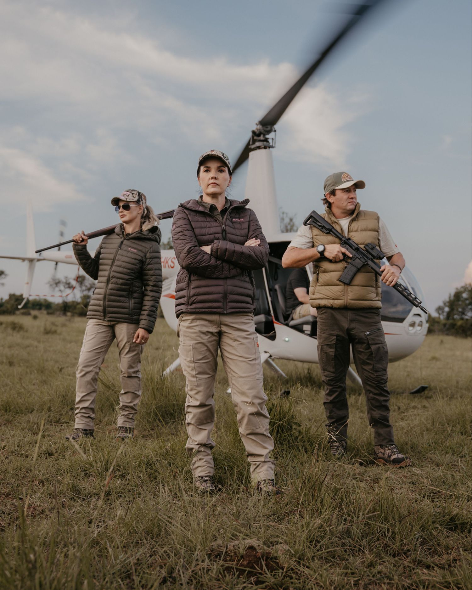 Three hunters standing in a field with a helicopter in the background on an overcast day.