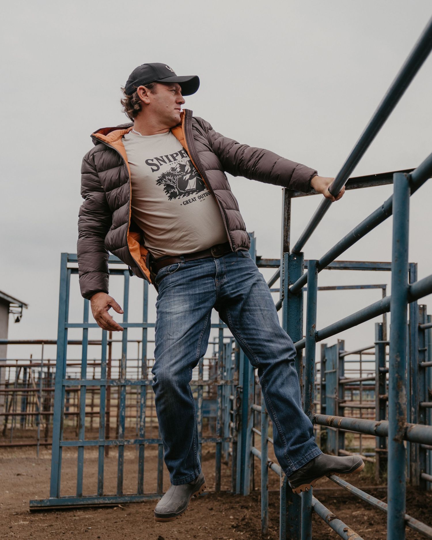 Man in puffer jacket and cap leaning on metal fence at a ranch livestock pen.