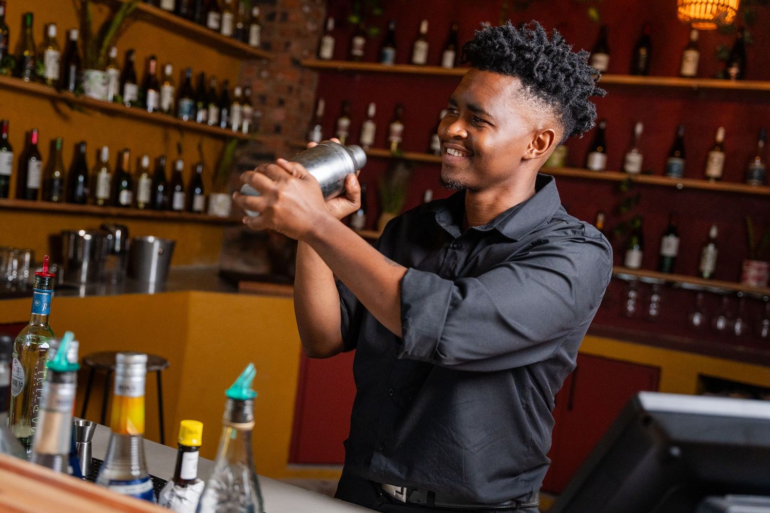 Smiling bartender shaking a cocktail mixer behind a well-stocked bar with bottles on shelves.