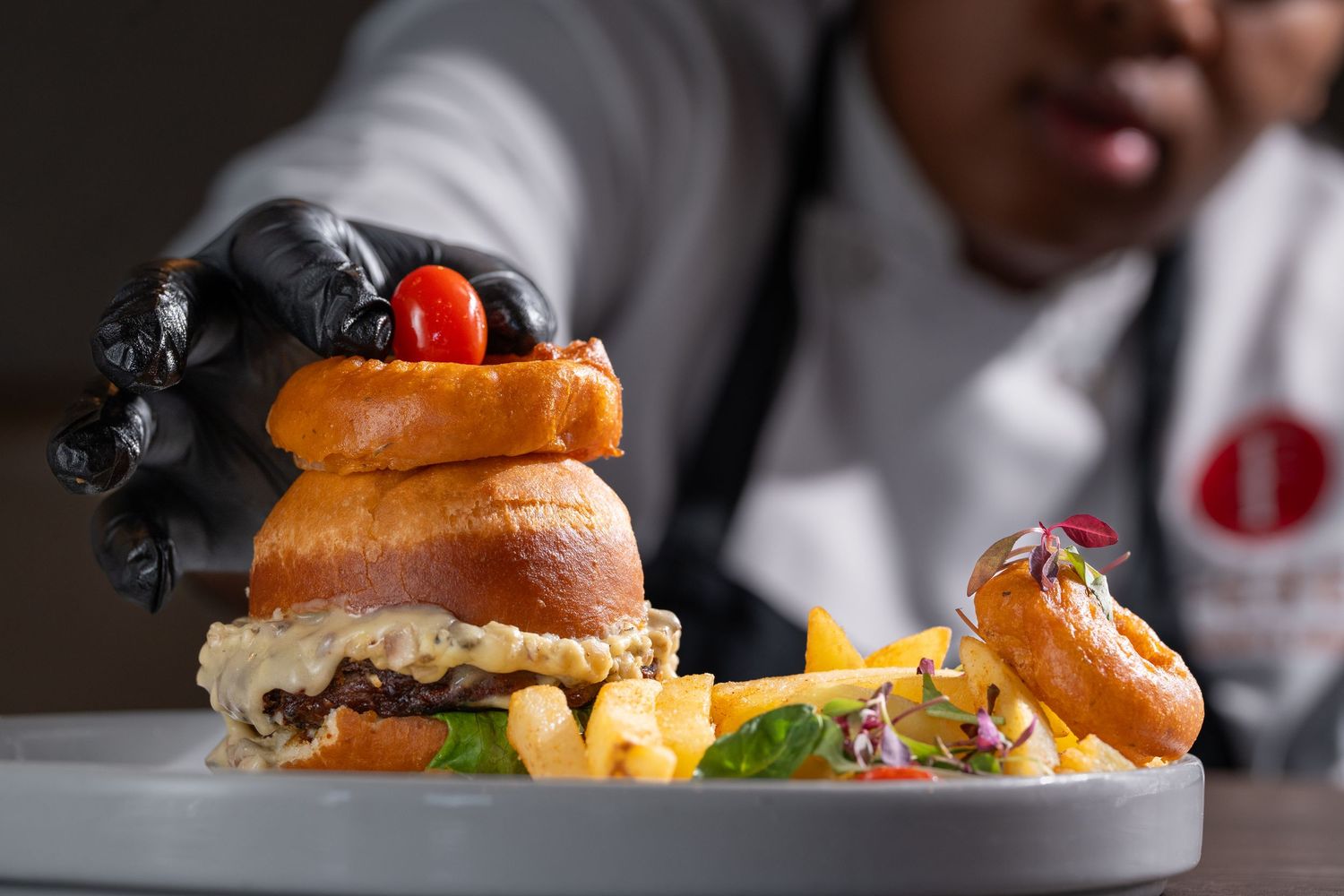 Chef presenting a towering gourmet burger with fries and a shrimp on a white plate.