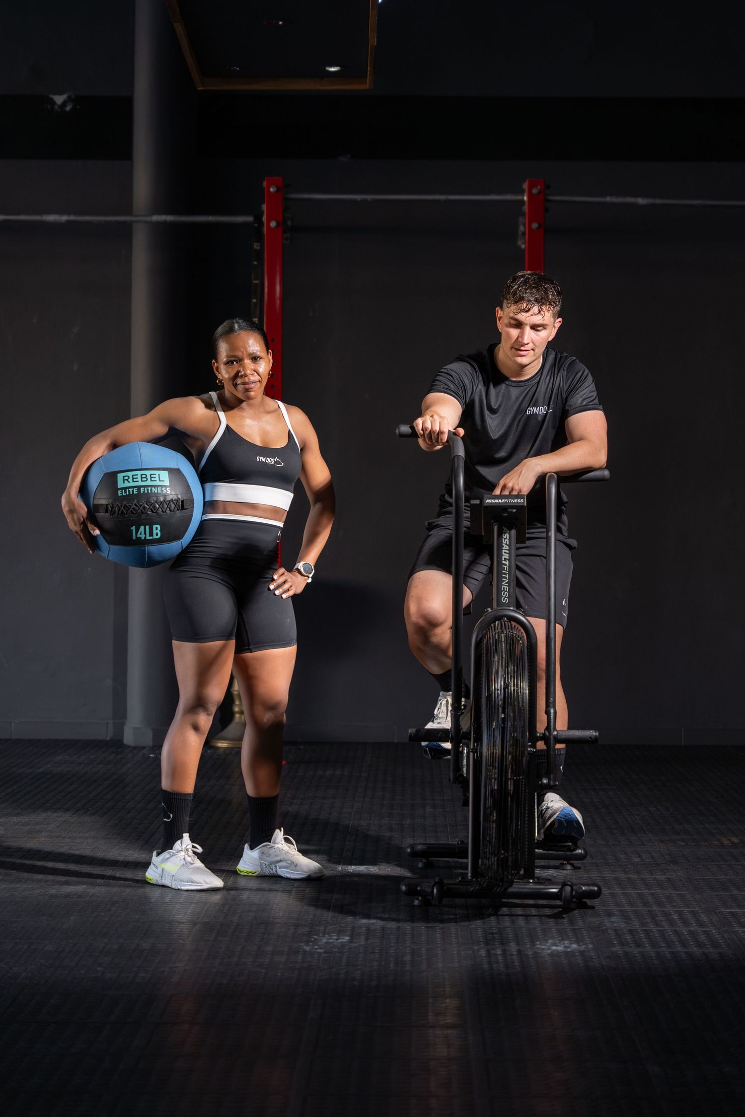 Man and woman posing with gym equipment including a 14lb medicine ball and air bike in dark fitness studio.