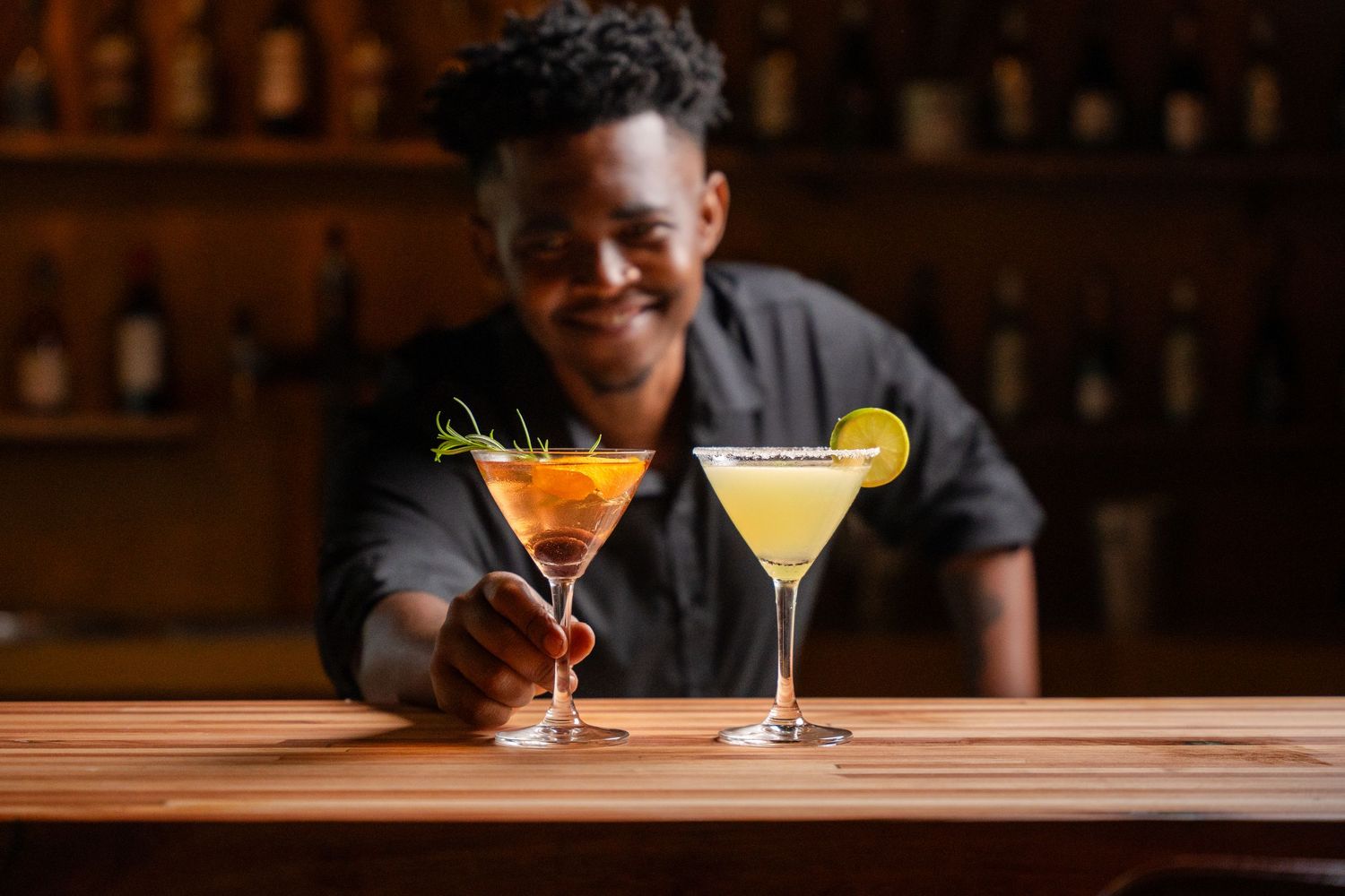 Smiling bartender presenting two colorful cocktails in martini glasses on a wooden bar counter.