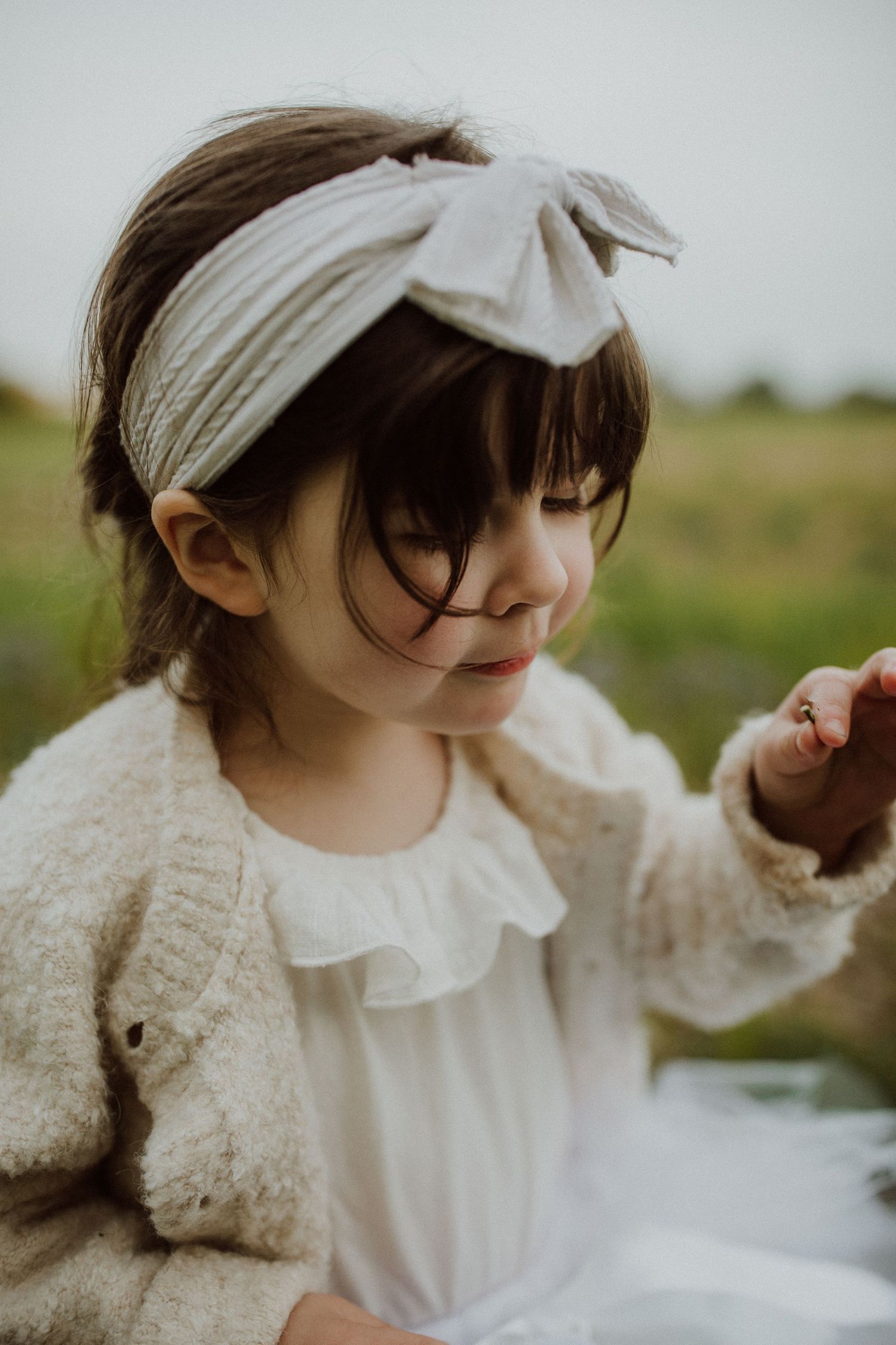 A dreamy outdoor portrait of a small child wearing a white bow headband and cozy cardigan in a natural setting.