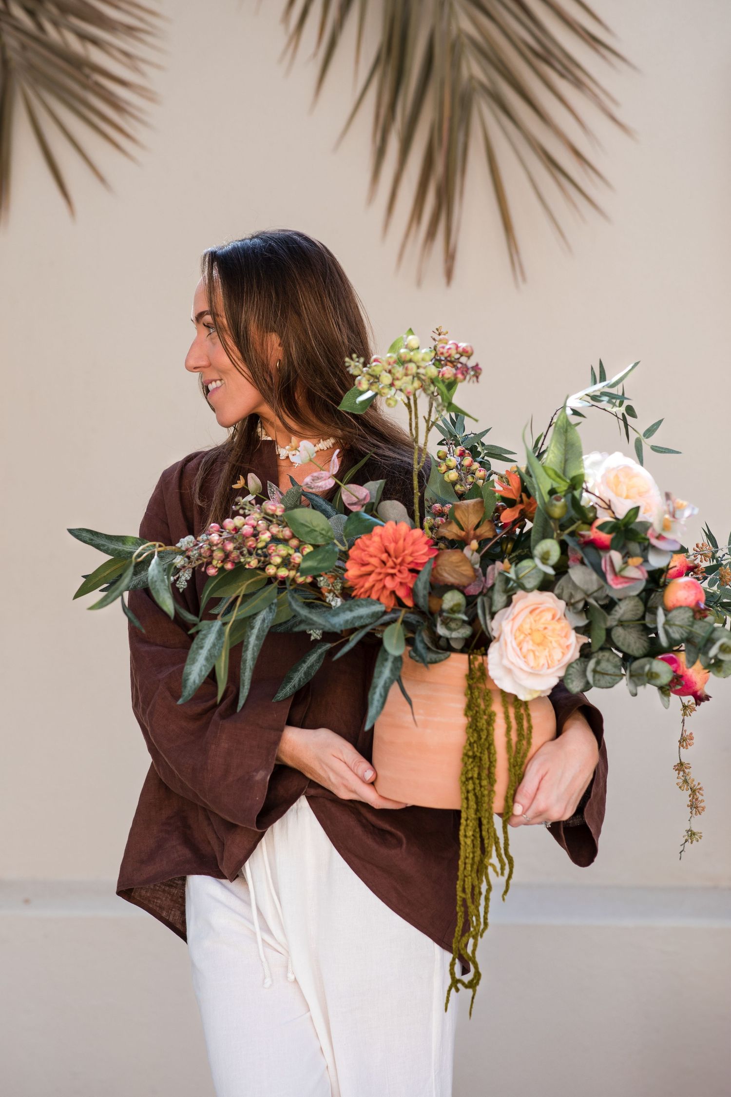 A florist in brown shirt carries a lush arrangement of coral and peach flowers with cascading greenery against a neutral wall.