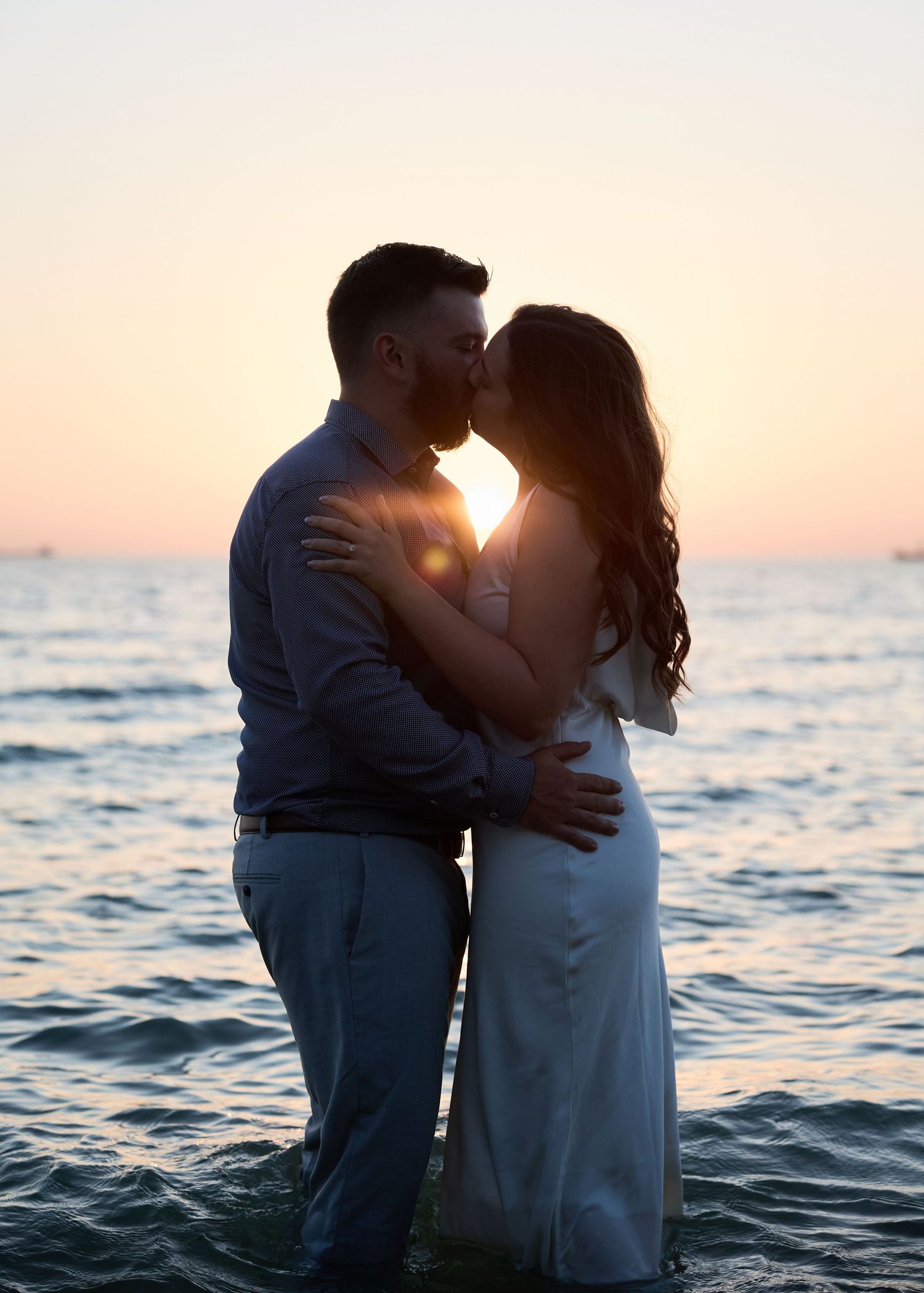 A romantic couple embraces on a beach at sunset with ocean waves in the background.