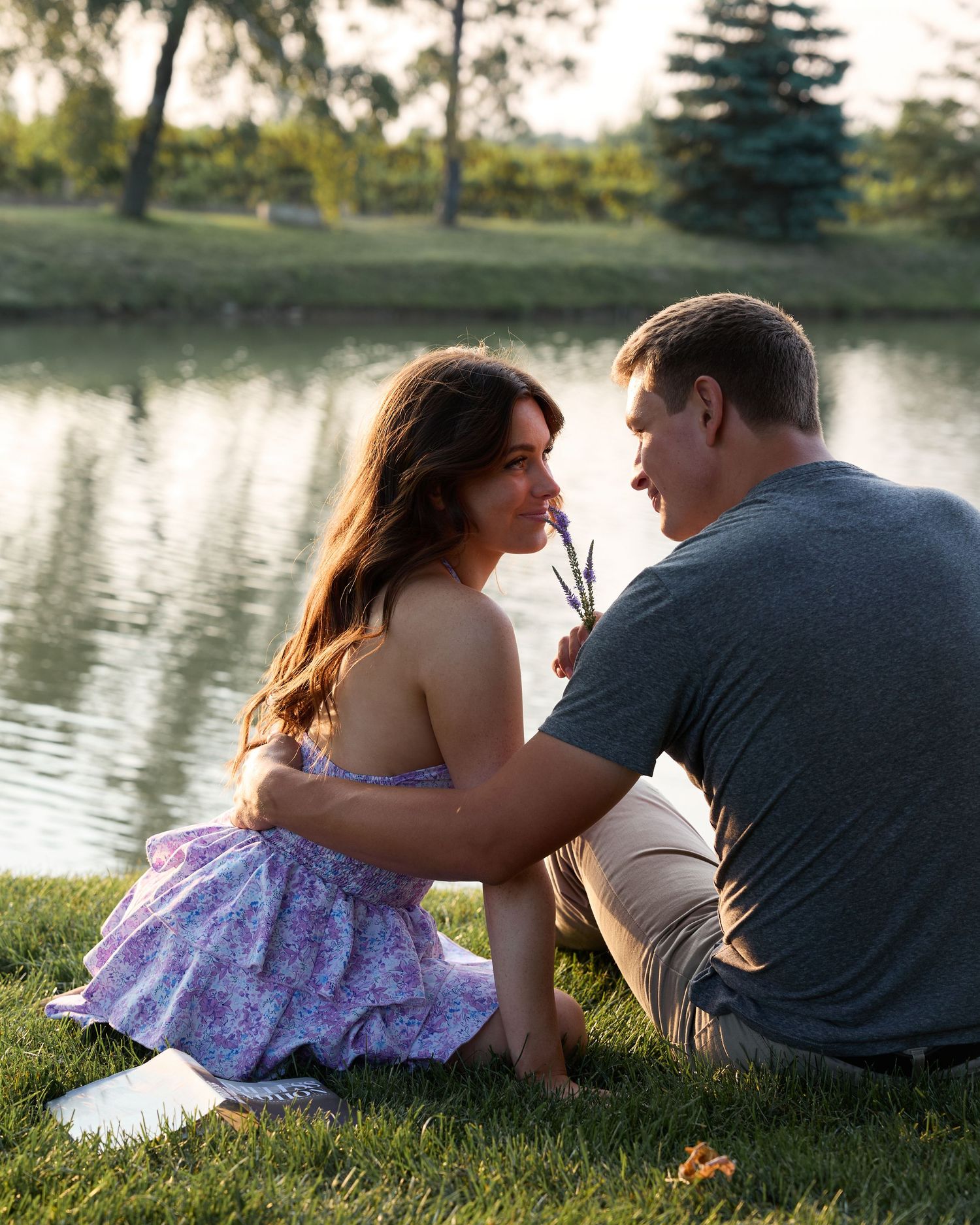 A couple relaxes on the grass beside a lake at sunset, sharing intimate moments in a romantic outdoor setting.