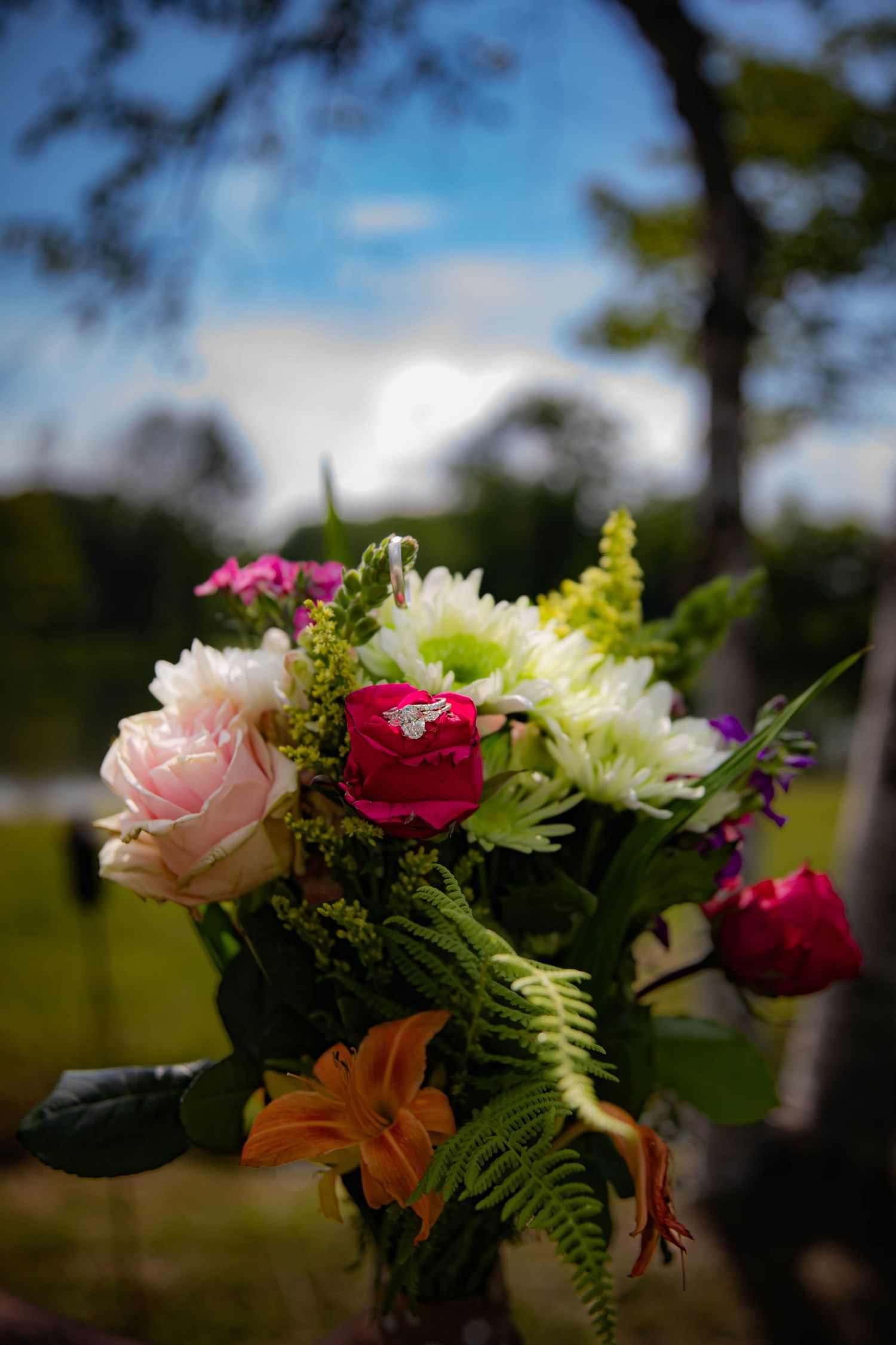 A vibrant bouquet featuring pink roses, red carnations, white daisies, orange lilies and ferns against a blurred outdoor background.