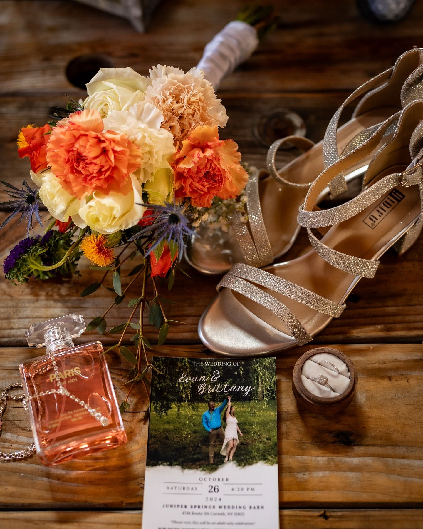 Wedding details flatlay with pink and peach floral bouquet, silver strappy heels, perfume bottle and invitation card on wooden surface.