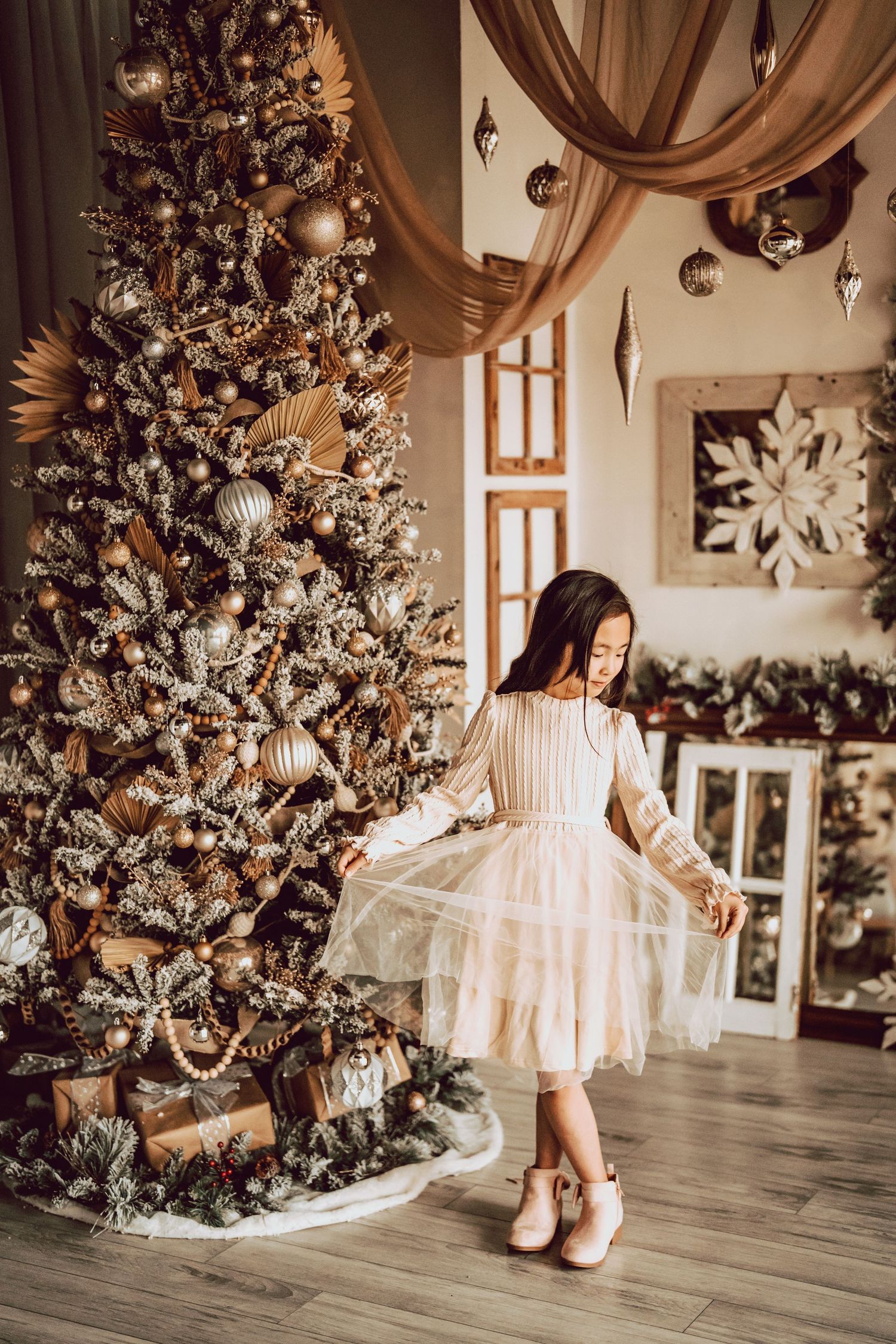 Young girl in white tulle dress twirling beside decorated gold Christmas tree with wrapped gifts during holiday photo session