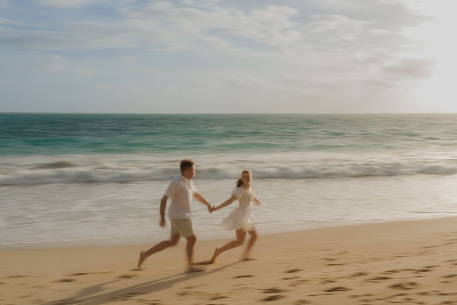 Couple running hand in hand along a sunny beach with turquoise ocean waves behind them.