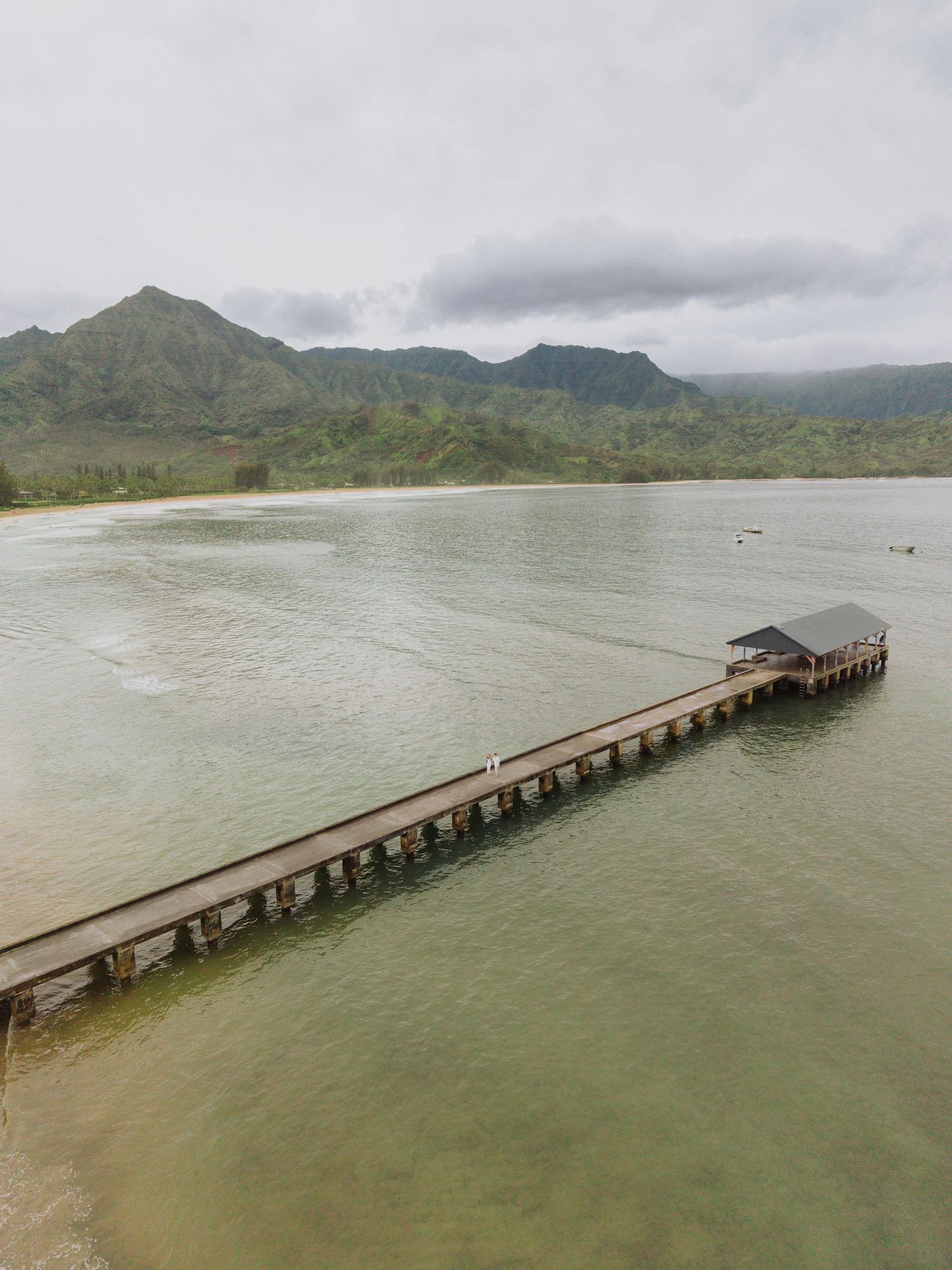 Aerial view of a wooden pier extending into calm Hawaiian bay with mountain backdrop.