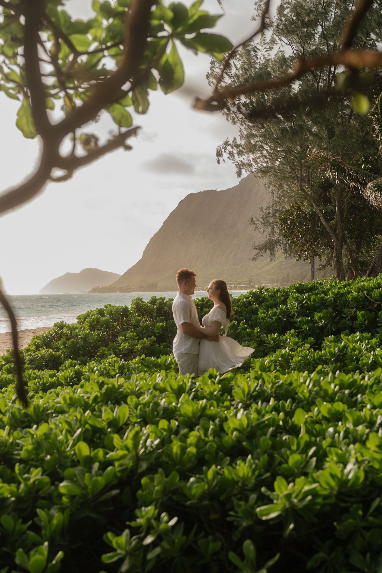 Couple embracing among lush tropical greenery on a Hawaiian beach with mountain views.