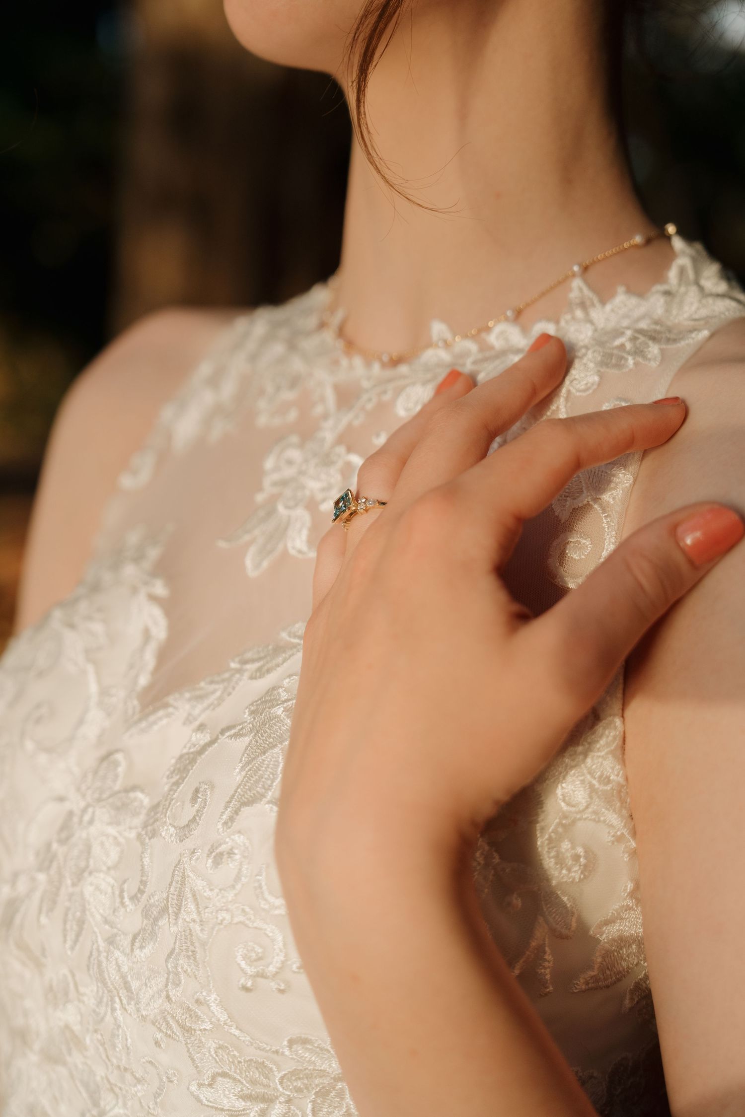 Bride in lace wedding gown touching chest wearing green gemstone ring and delicate gold necklace.