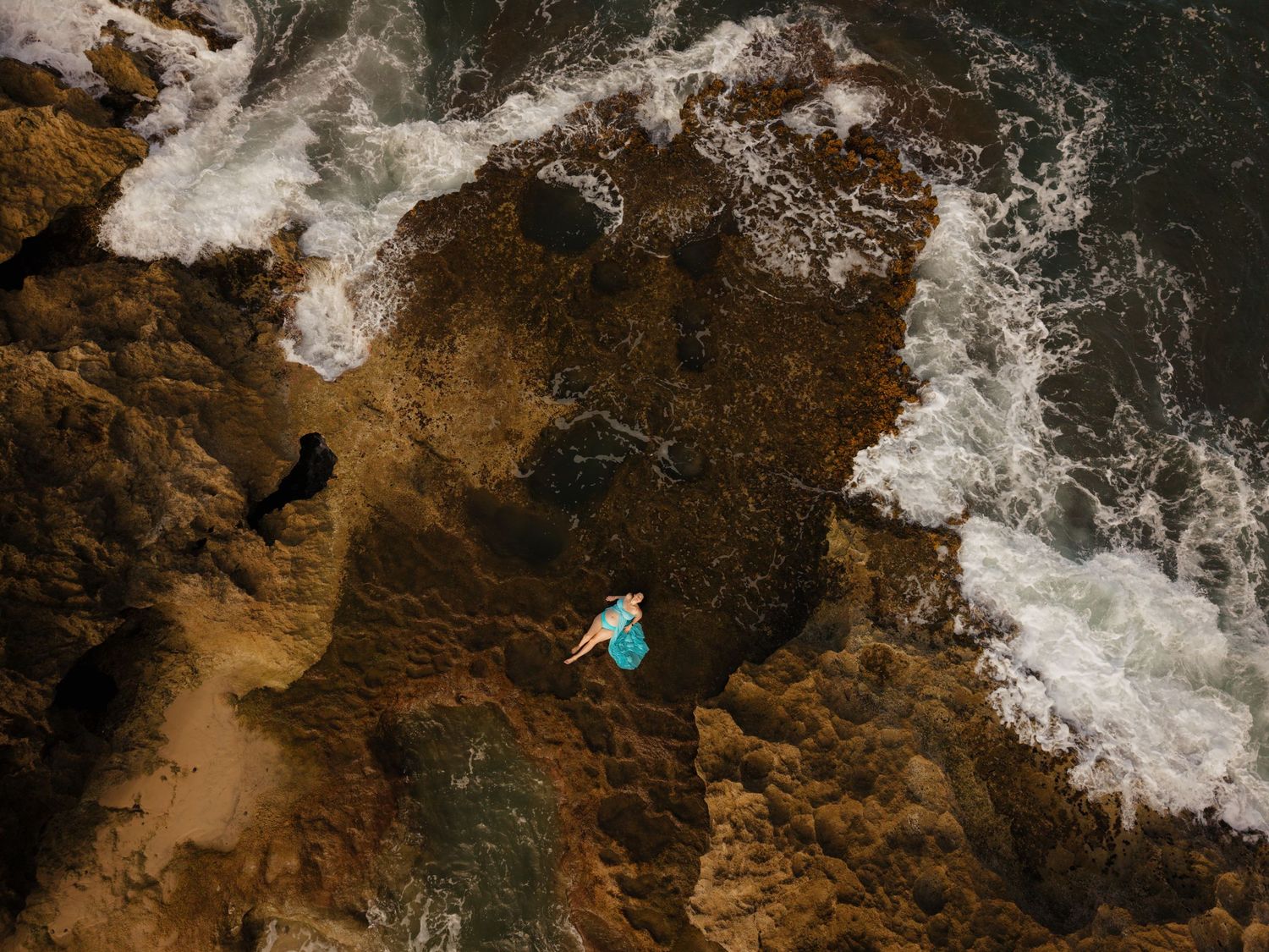Aerial view of a person lying on rocky coastal cliffs surrounded by crashing ocean waves.