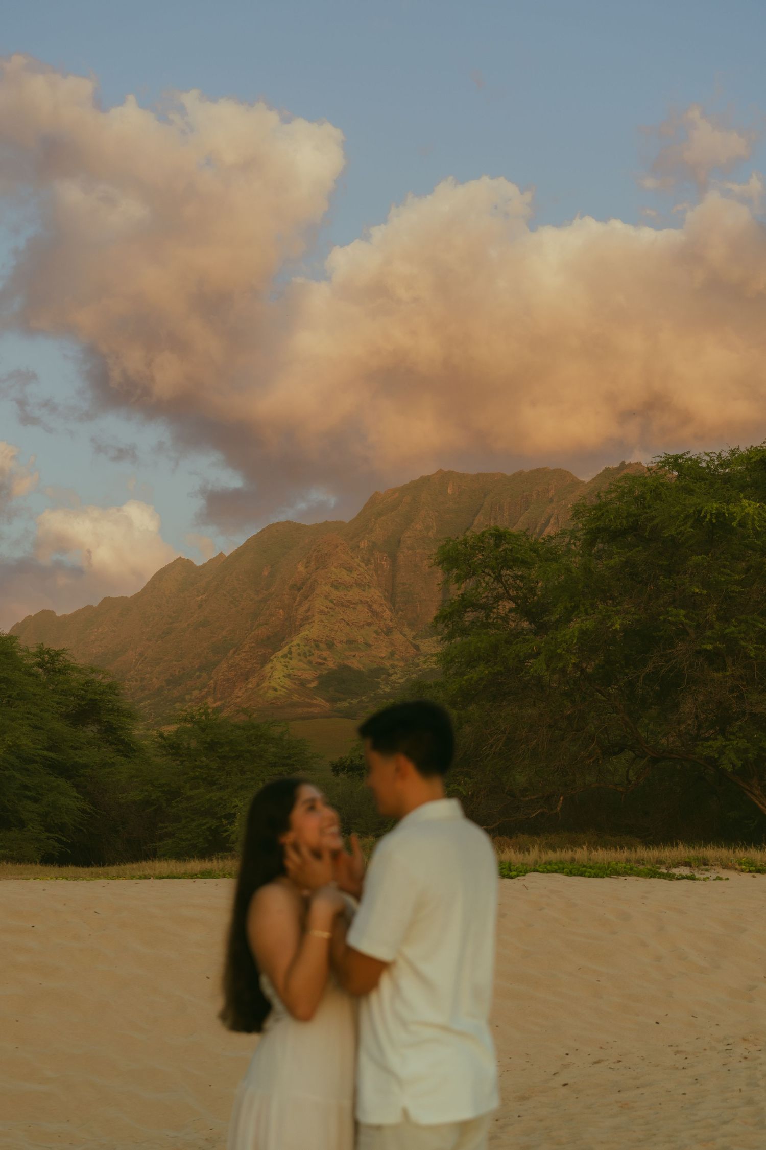 Couple in white embracing on sandy beach at sunset with lush mountains and pink clouds in Hawaii.
