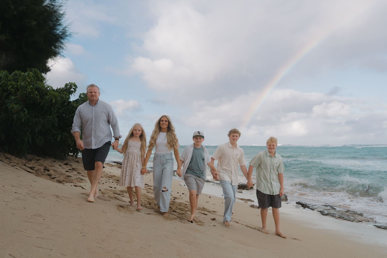 Family of six walking hand in hand on sandy beach with rainbow over ocean backdrop.