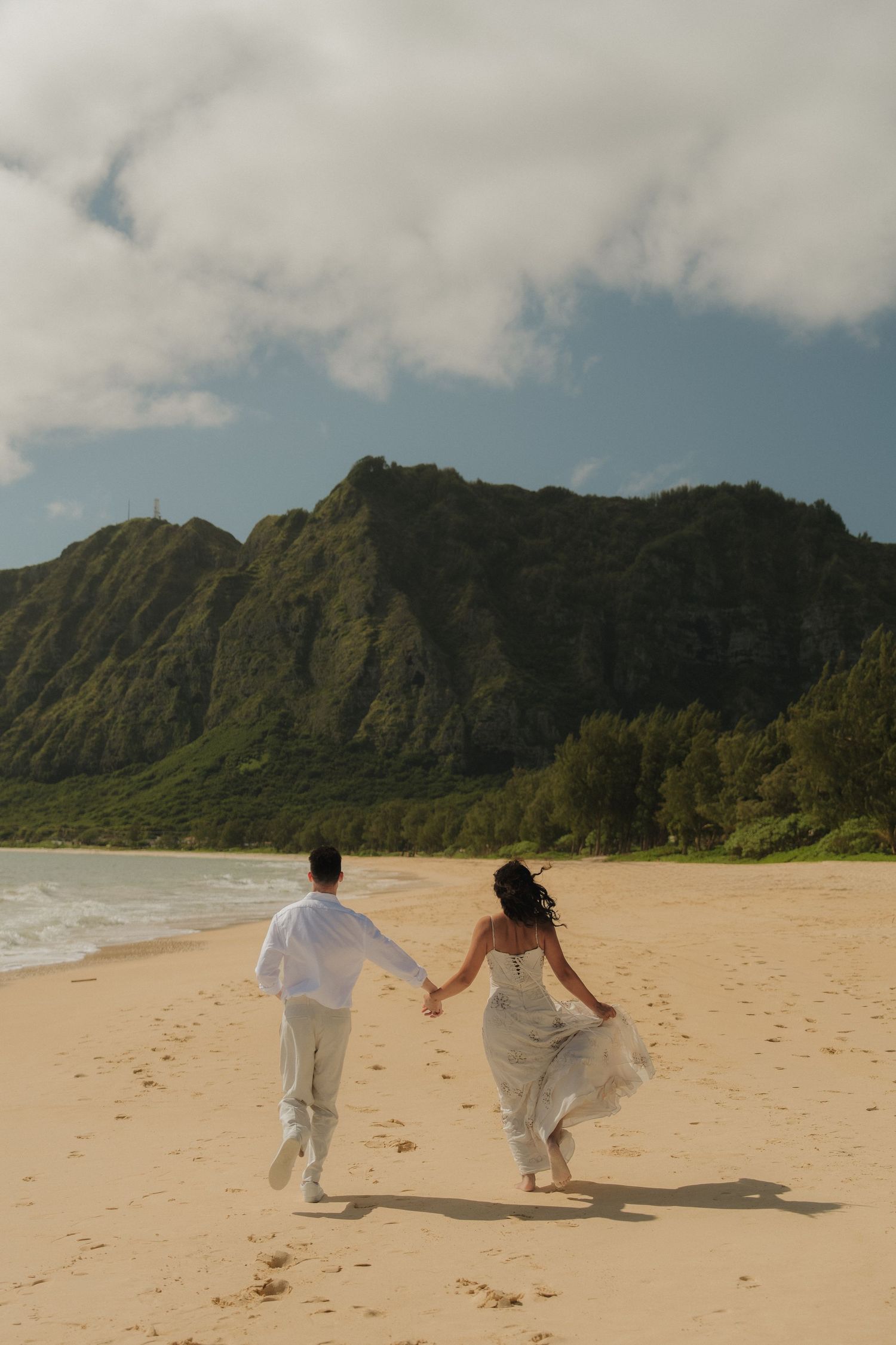 Couple in white holding hands running along a sandy Hawaiian beach toward lush green mountains.