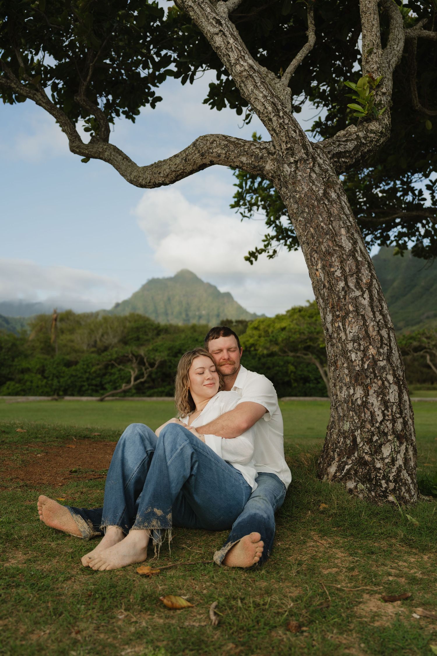 Couple sitting peacefully under a large tree with Hawaiian mountains and blue sky backdrop.