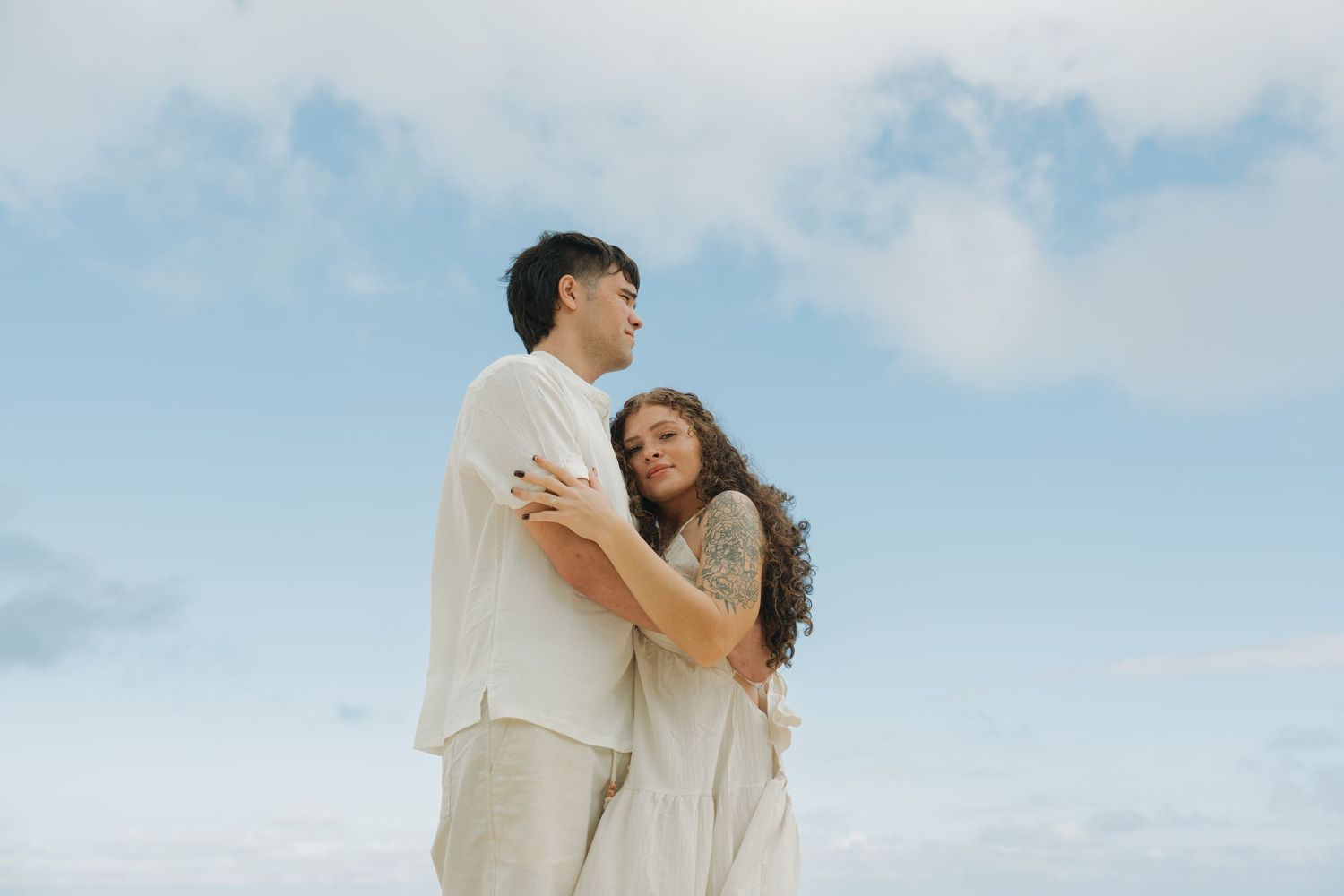 A couple embraces tenderly against a cloudy blue sky, wearing neutral linen outfits in a romantic outdoor portrait.
