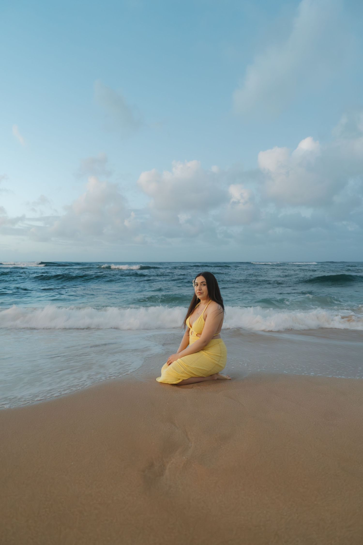 Woman in yellow dress kneeling on sandy beach with ocean waves and cloudy sky behind her.