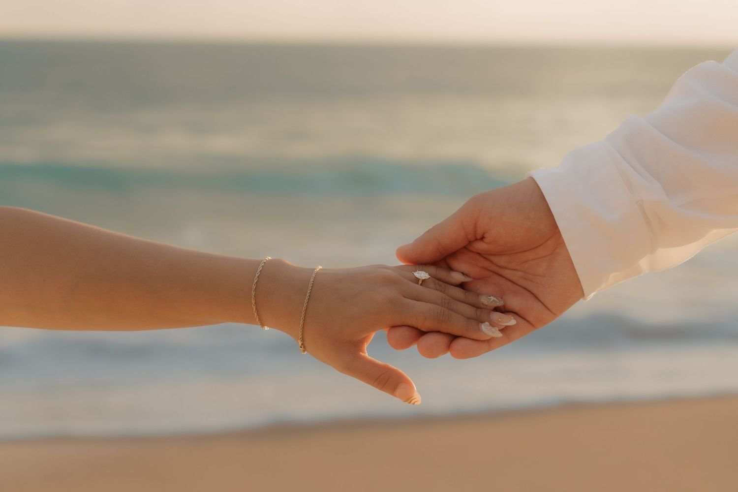 Close-up of couple holding hands on a beach, woman wearing a delicate gold bracelet at sunset.